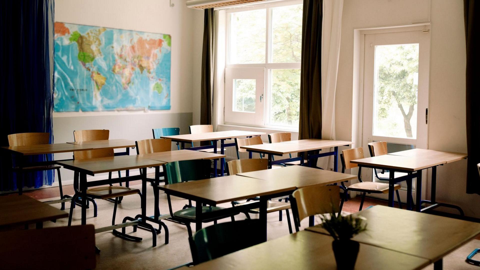 Photo of empty classroom with map of the world on the wall at the back. The classroom is tidy and all the chairs are tucked into the tables. There are large windows with trees showing on the far side.