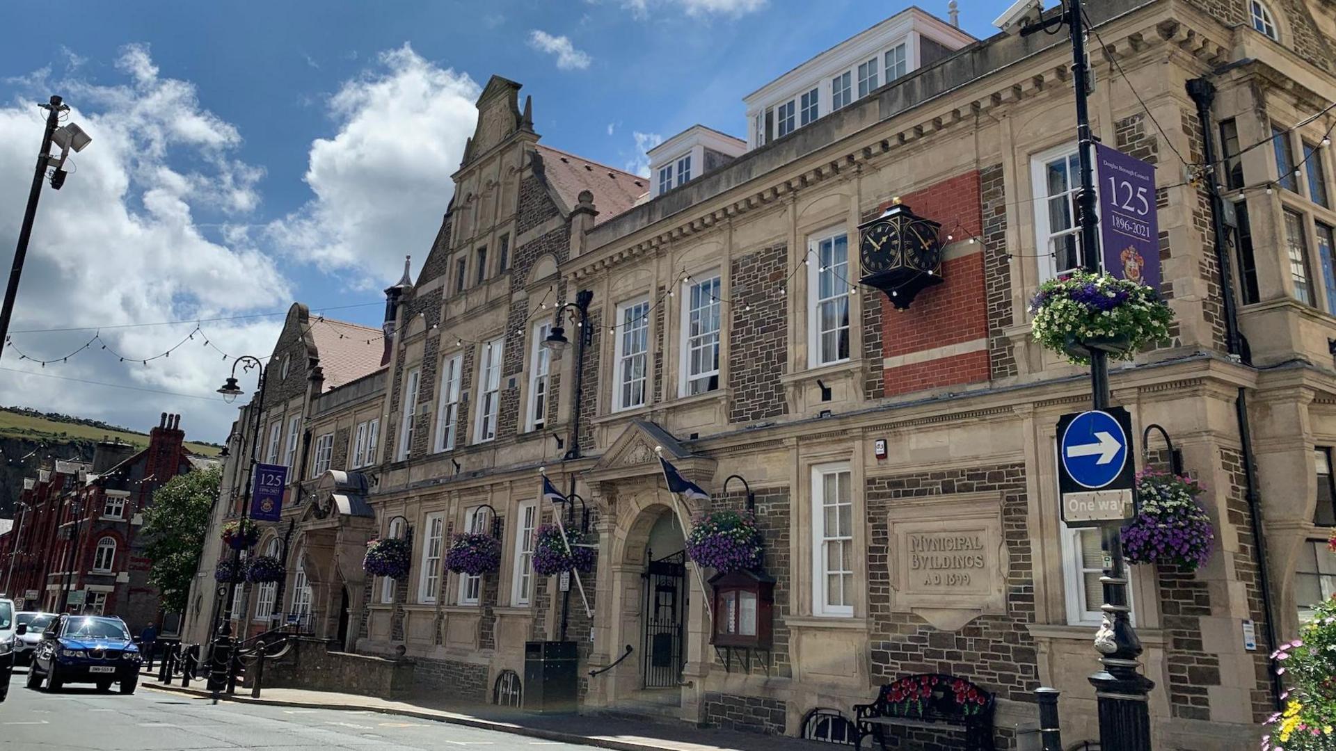 A view of the outside of the Douglas Council buildings on a sunny day. It has an ornate frontage and is decorated with hanging baskets.