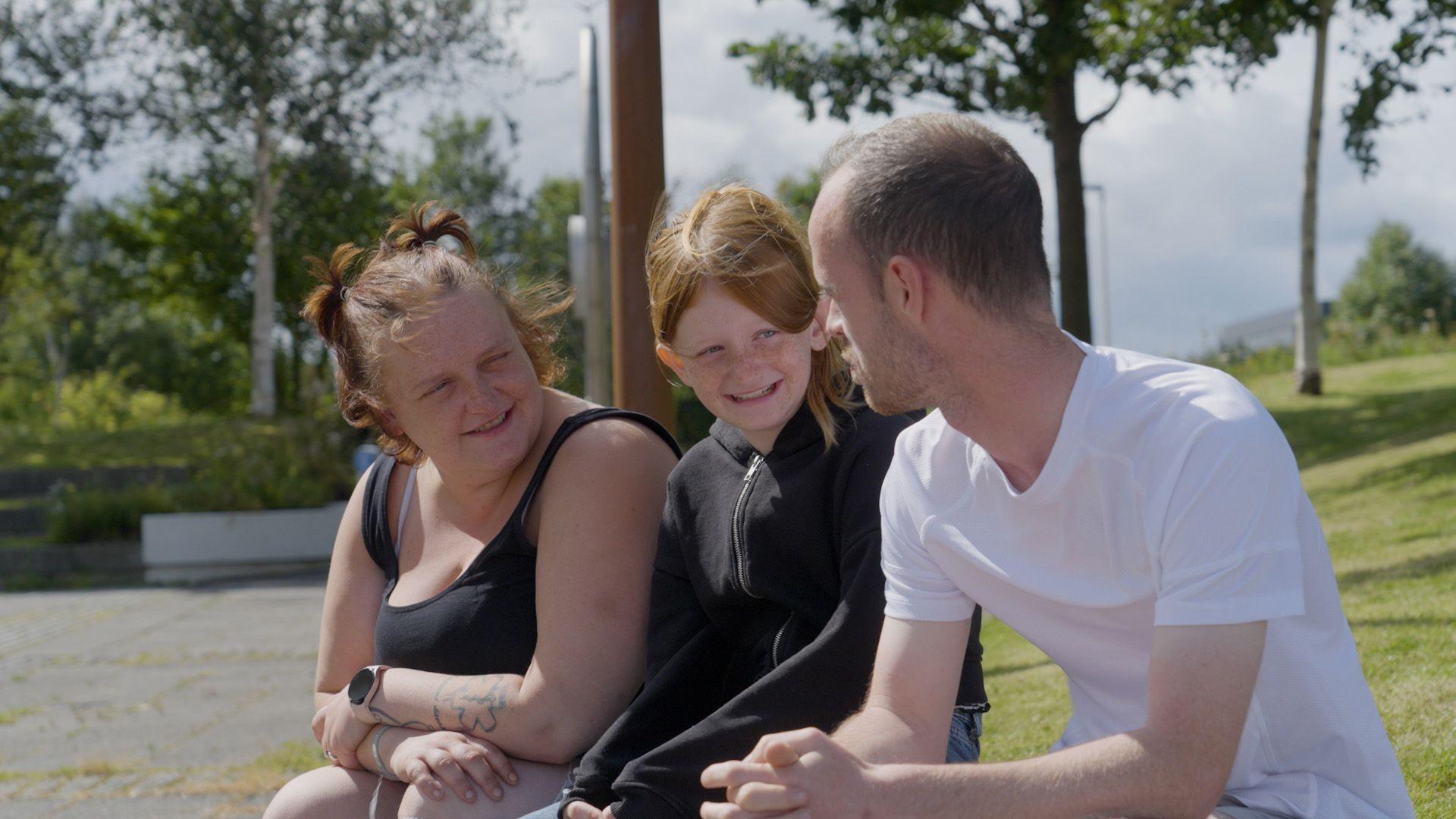 A young girl wearing back and with red hair, sits in-between her parents while sitting outside on a sunny day. They are all looking smiling at each other. The mother has auburn hair and a black top. The dad has close-cropped hair and a white T-shirt.