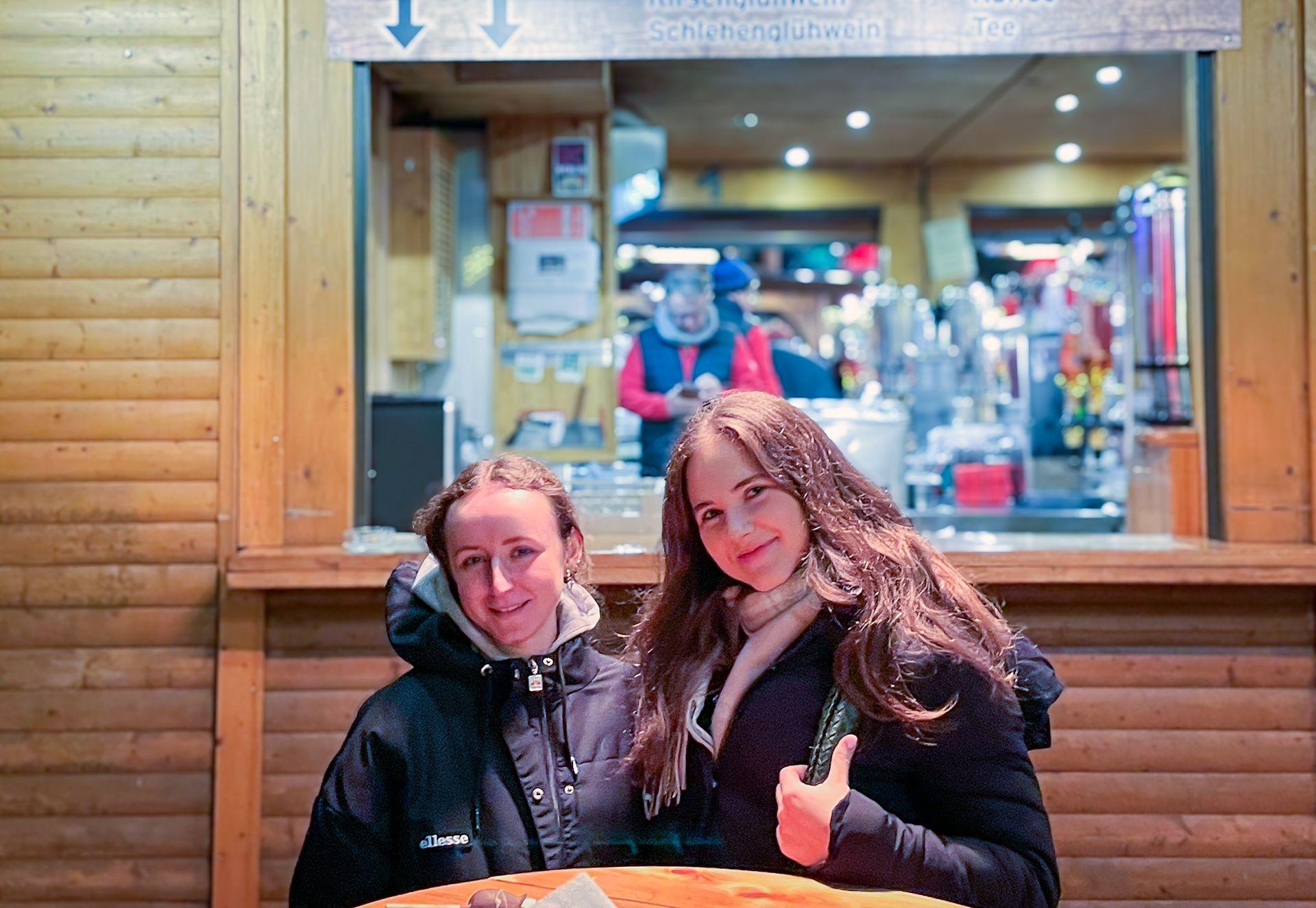 Two young women sit at a wooden table in front of a wooden hut selling chocolate covered strawberries at a Christmas market in Berlin. They are both smiling.