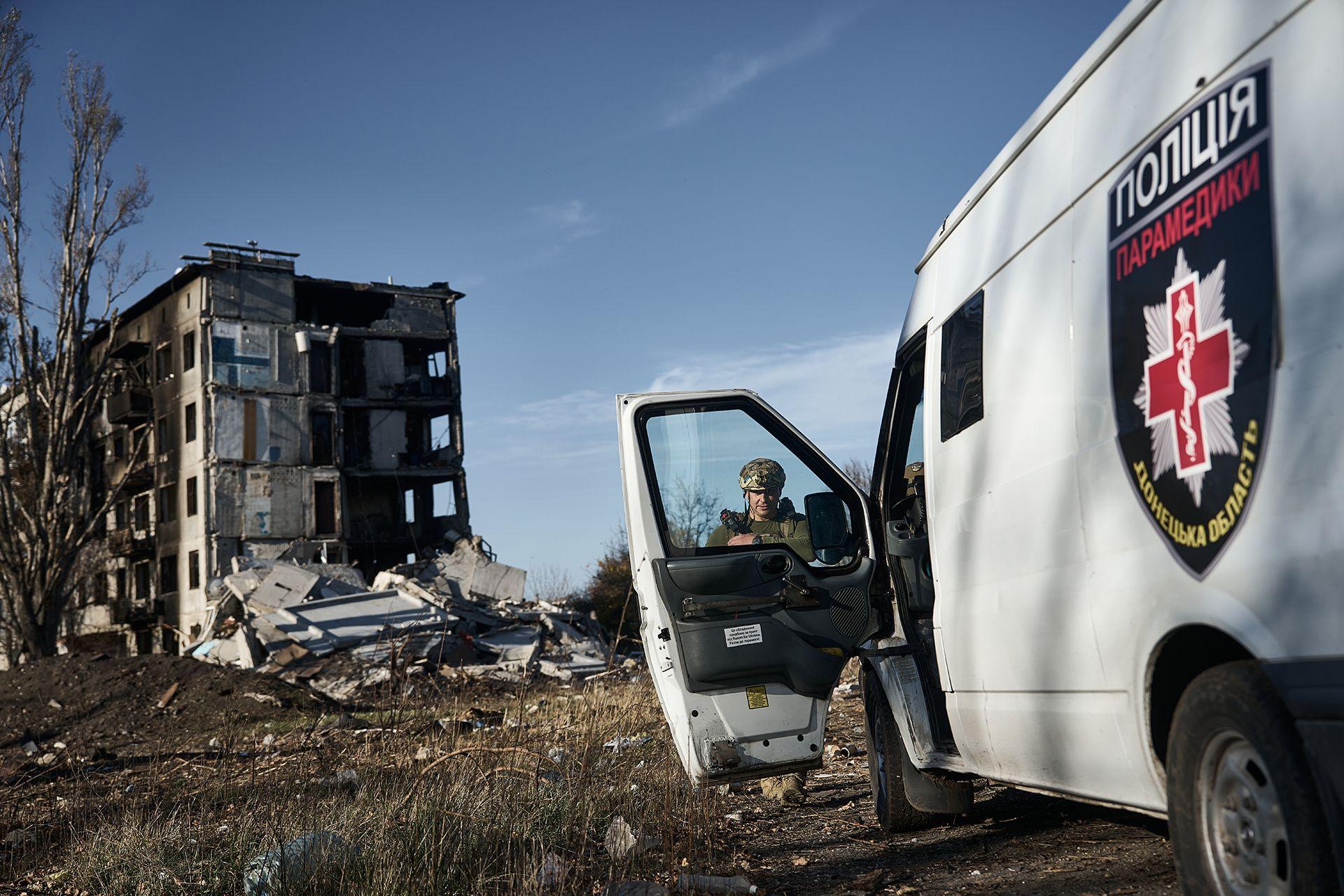 A police officer dressed in camouflage is just seen through the window of an open door of a police van belonging to the 'White Angels' - he's looking down to the floor and holding his weapon, and in the backtground is a burnt out building with rubble next to it against a blue sky, in Avdiivka, eastern Ukraine on 30 October 2023.