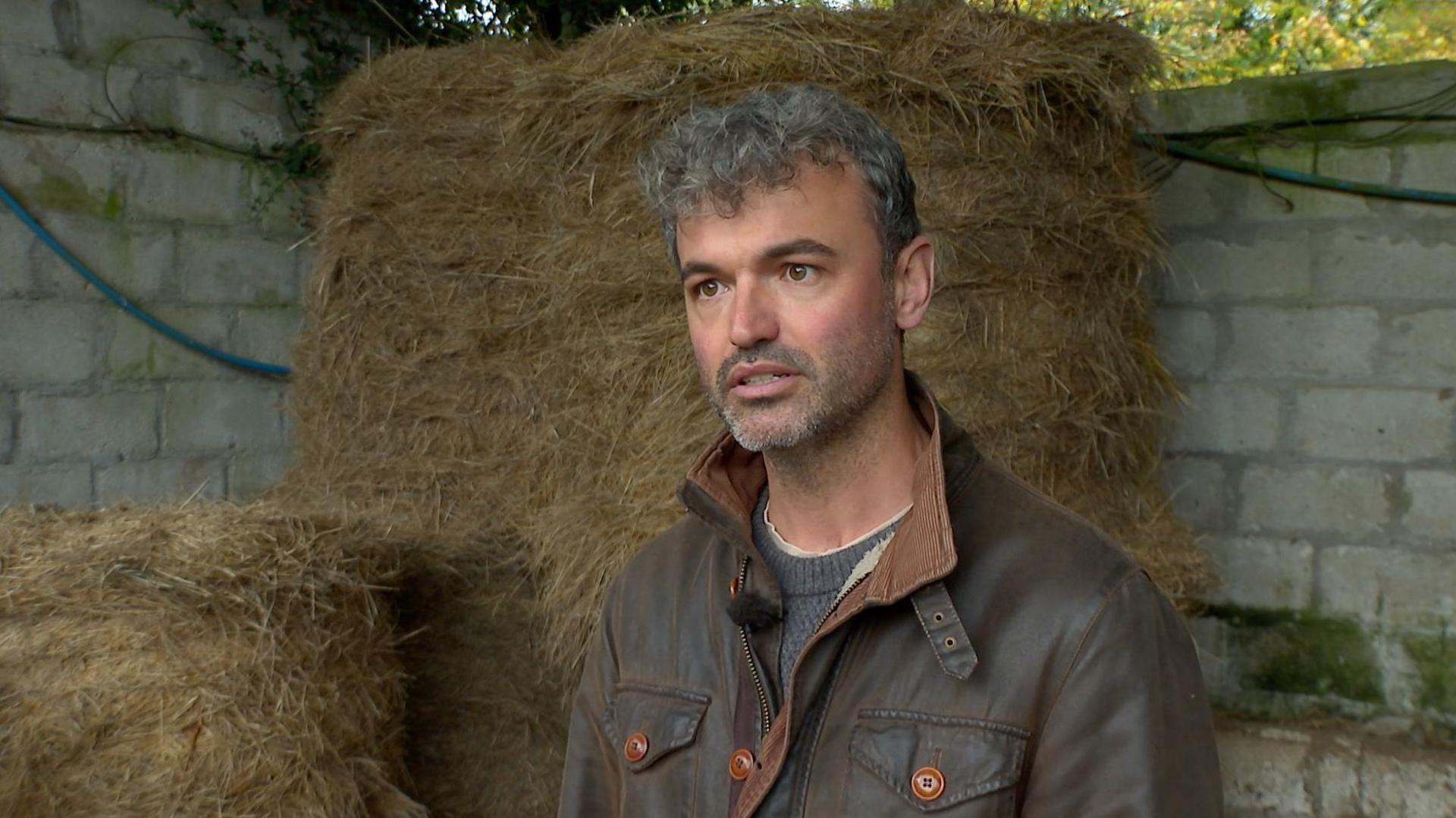 Stuart Oates facing the camera. He is wearing a brown leather jacket, with a grey knitted sweater underneath. He has grey hair and a small beard. Behind him is a stack on hay bales wrapped in sisal twine. Behind the hay is a grey brick wall.