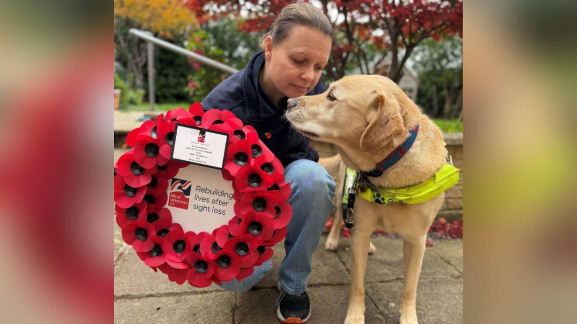 Kelly in a garden, crouching down beside her guide dog Archie who has a harness on and his head is turned towards Kelly. She is holding a large wreath of poppies which says within the circle 'Rebuilding lives after sight loss'