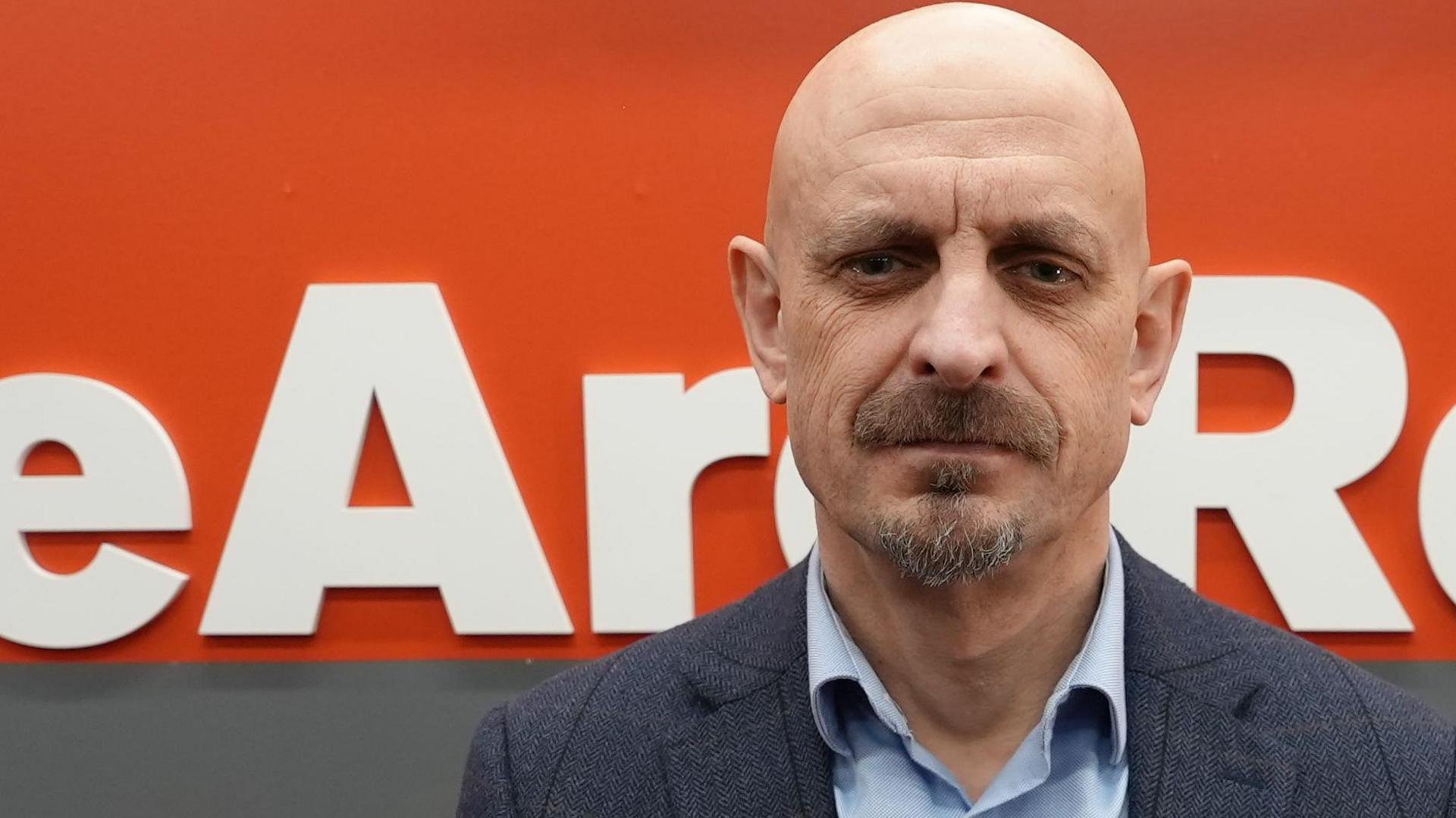 Dave Perry. A man who has a moustache and is looking into the camera. He is dressed in a blue shirt and suit jacket. He is sat down and behind him is a grey and orange backdrop. On the orange backdrop it says WeAreRebels