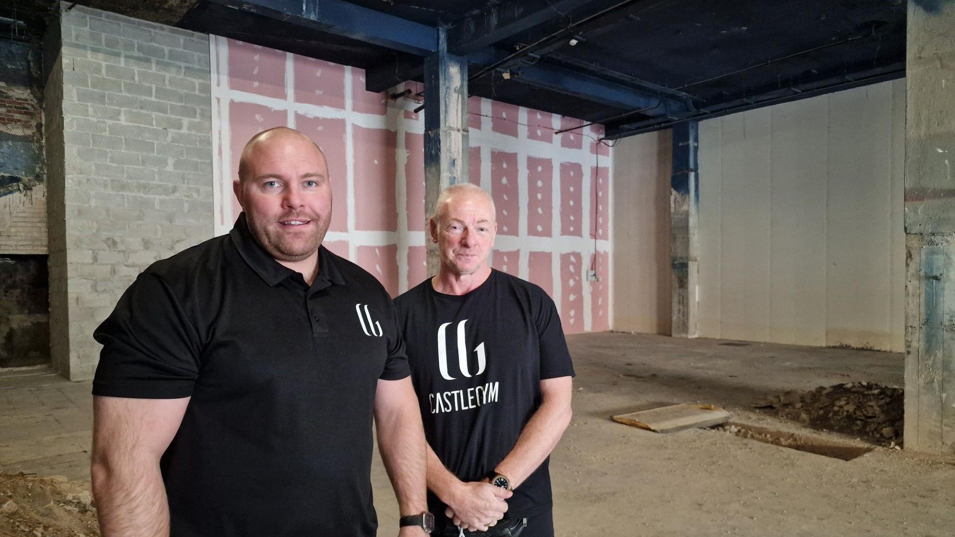 Two bald men in black gym-branded t shirts standing inside a building in need of some work itself