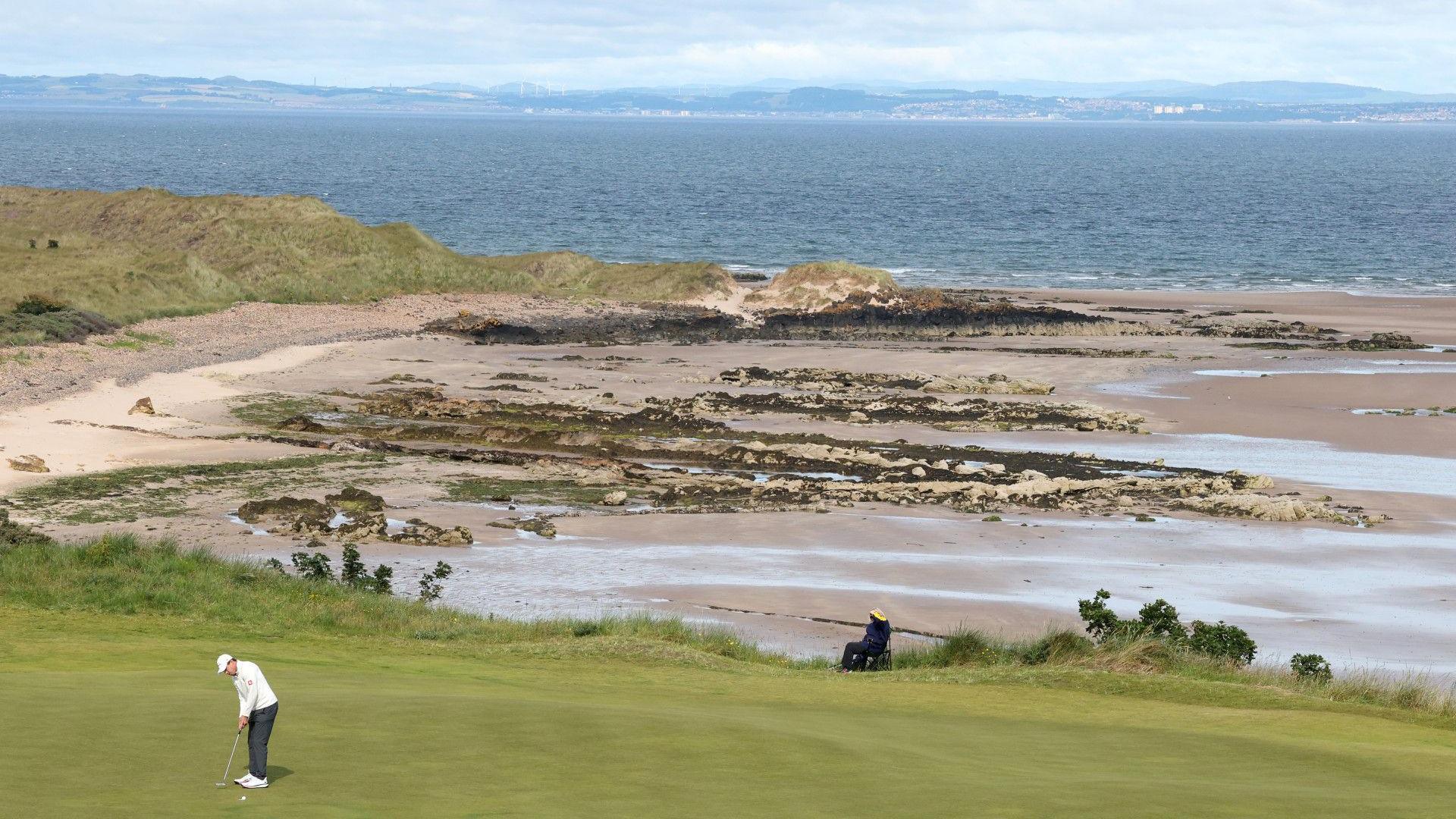 Matt Fitzpatrick of England putts on the 13th green during the pro-am prior with the beach and the North Sea in the background. He is wearing a white cap and jumper, grey trousers and white golf shoes. He is a small figure in the bottom left of the picture, which focuses on the background scenery