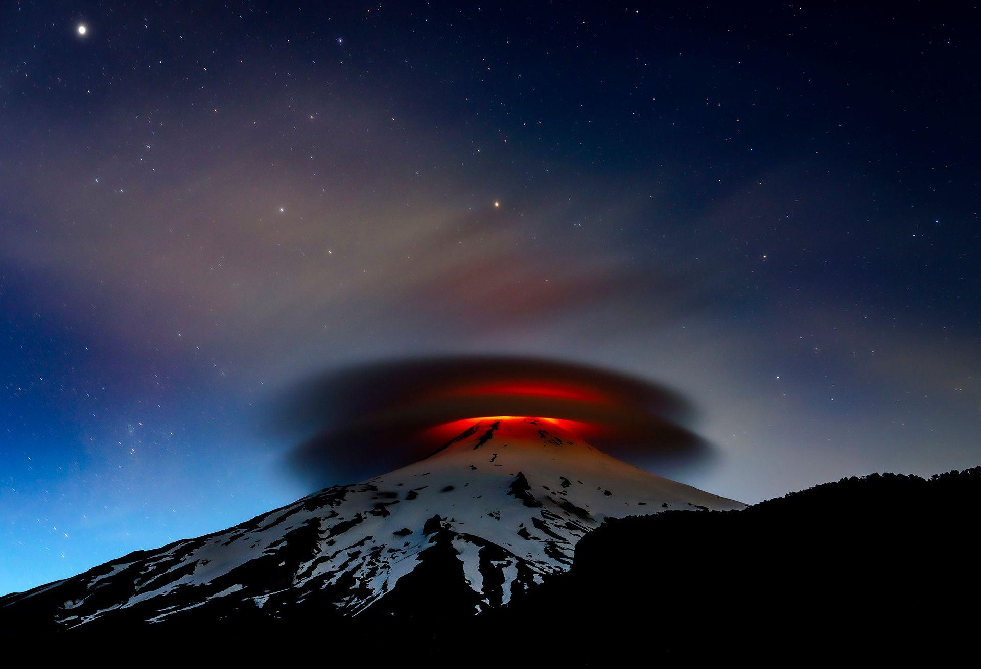 A fiery red glow from Chile’s Villarrica volcano illuminates the night sky. Above it, a lenticular cloud takes on an eerie, swirling shape, glowing in the reflected light.