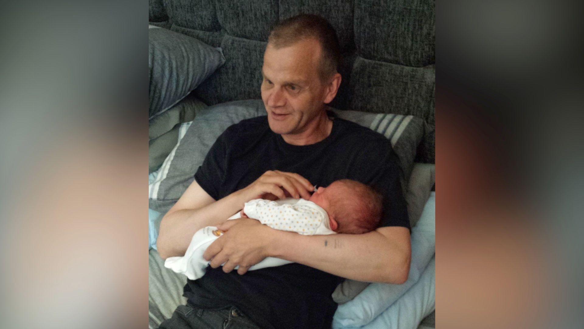 A man wearing a black t-shirt leaning back on a grey bed holding a baby in his arms.