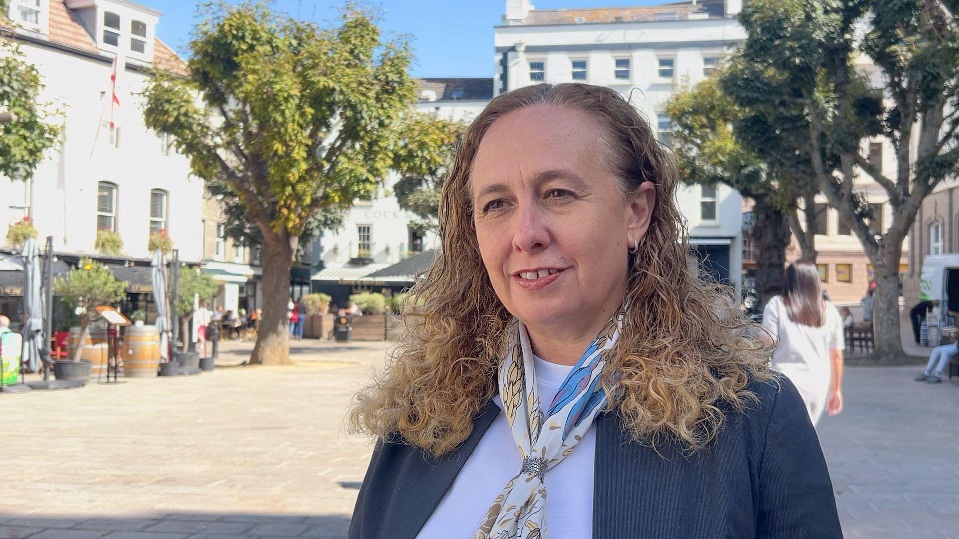 Deputy Inna Gardiner is wearing a dark blue blazer and a white T-shirt. She has a small scarf on and is standing in Jersey's Royal Square. 