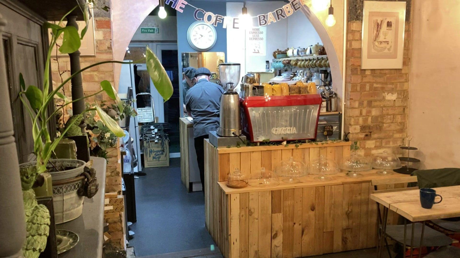 Daren Wayland works in his coffee shop with his back to the camera. A counter with empty cake containers and a coffee machine with yellow mugs on top can be seen. Plants rest on a shelf close to the camera.