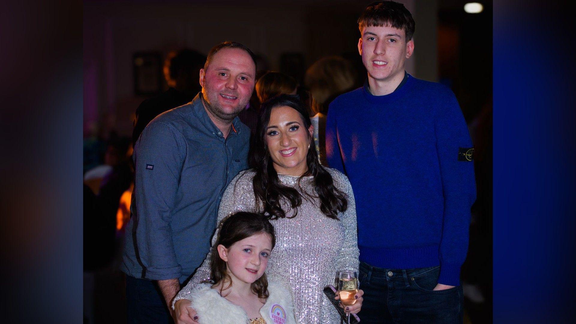 A family of four poses for a picture. The adult man on the left wears a blue shirt and the young man on the right - who is much taller than the others - wears a blue jumper. The adult woman stands in the middle with her arm around a young girl in a white fur coat.