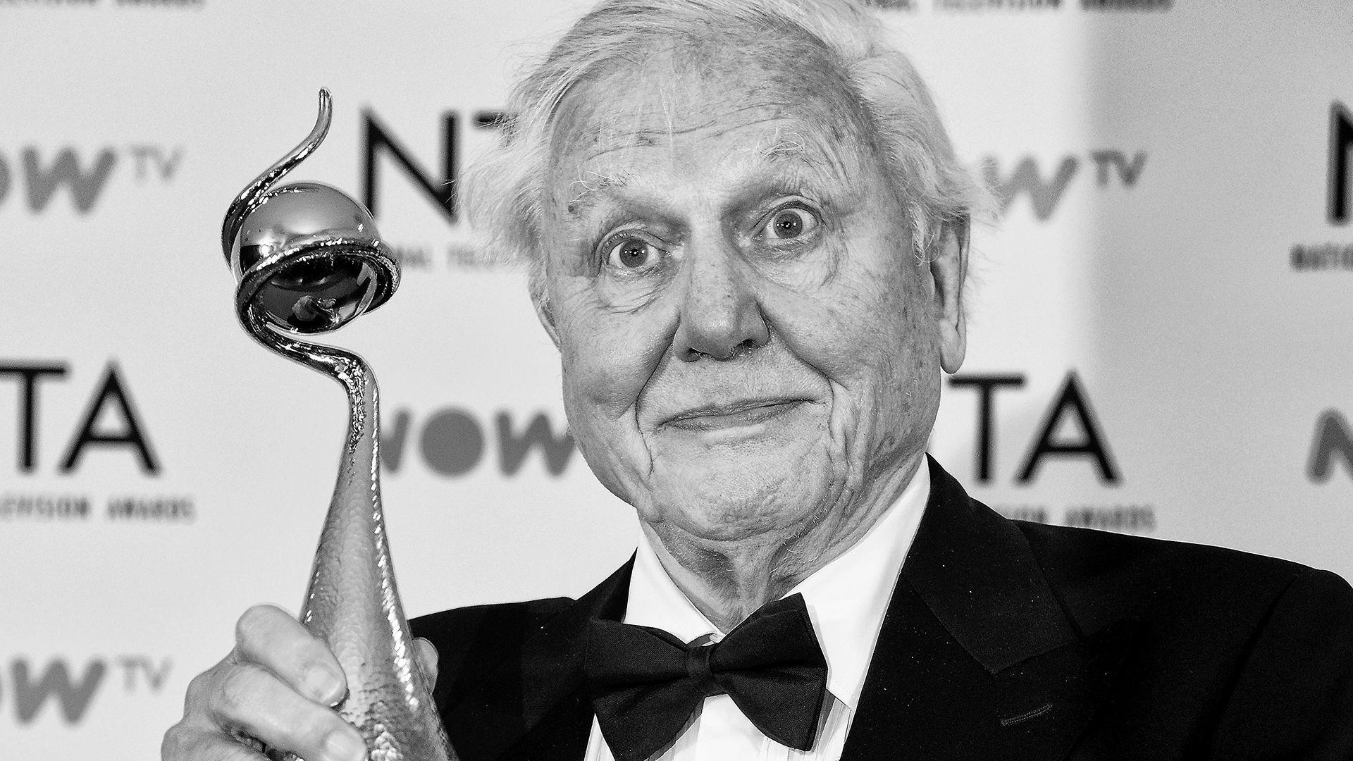 A black and white photo of Sir David Attenborough, an older man holding a trophy. 