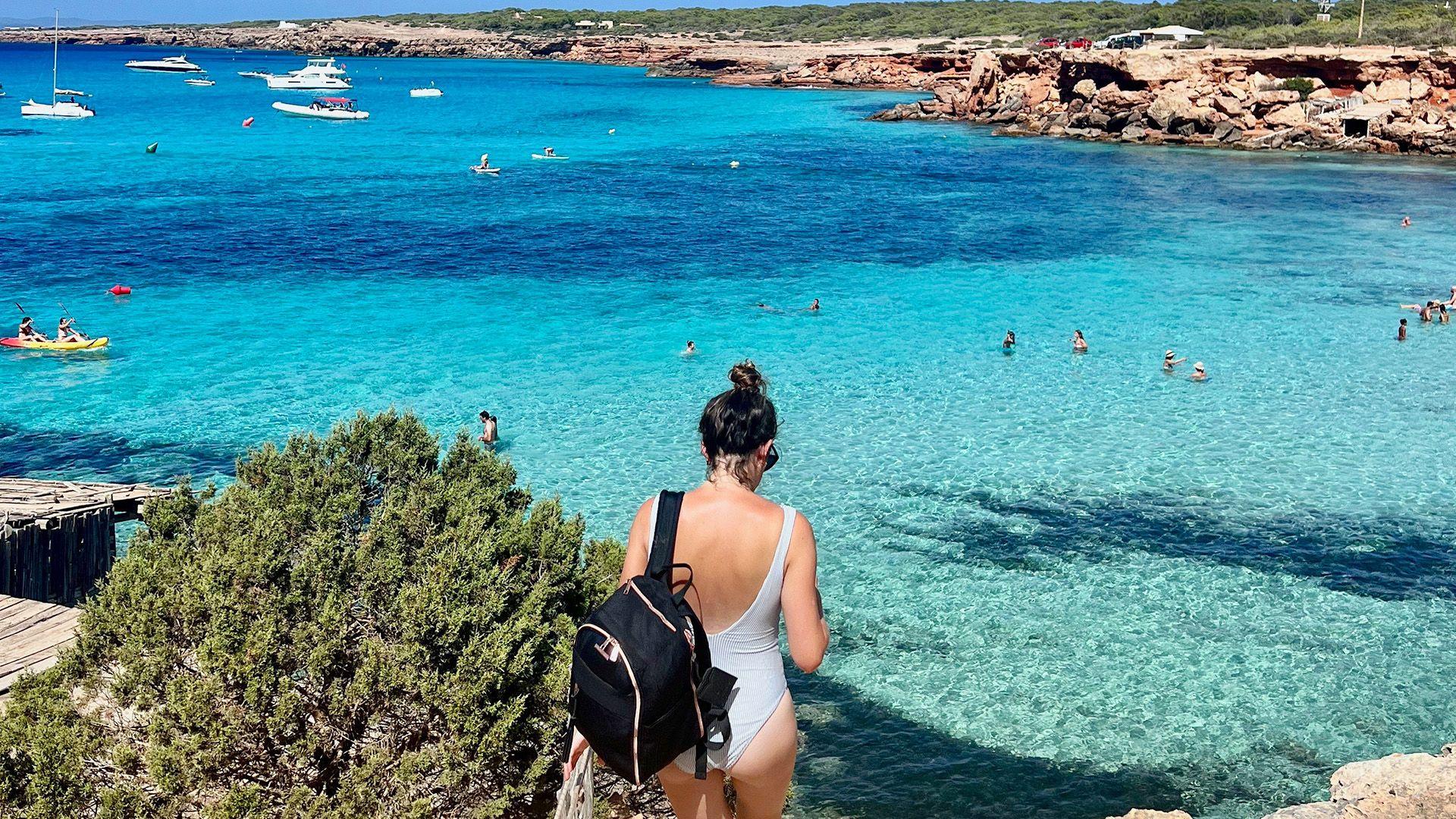 Monica is walking down to the beach in a swimsuit with a black backpack and a light blue swimsuit. Her back is facing the camera and ahead of her we can see bright blue waters. 