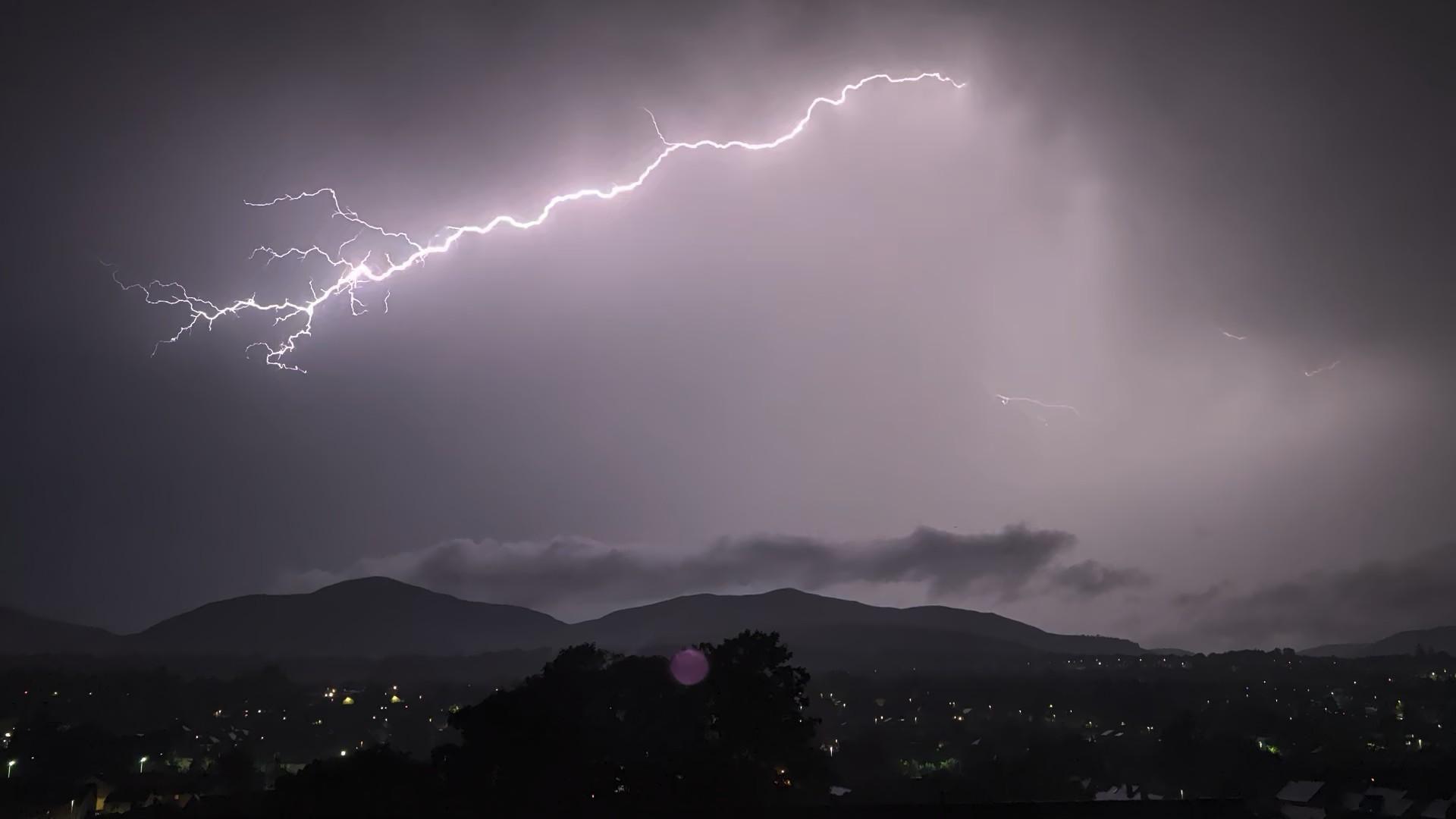 A dark night sky with the a bright lightning bolt. There are dark hills in the background.