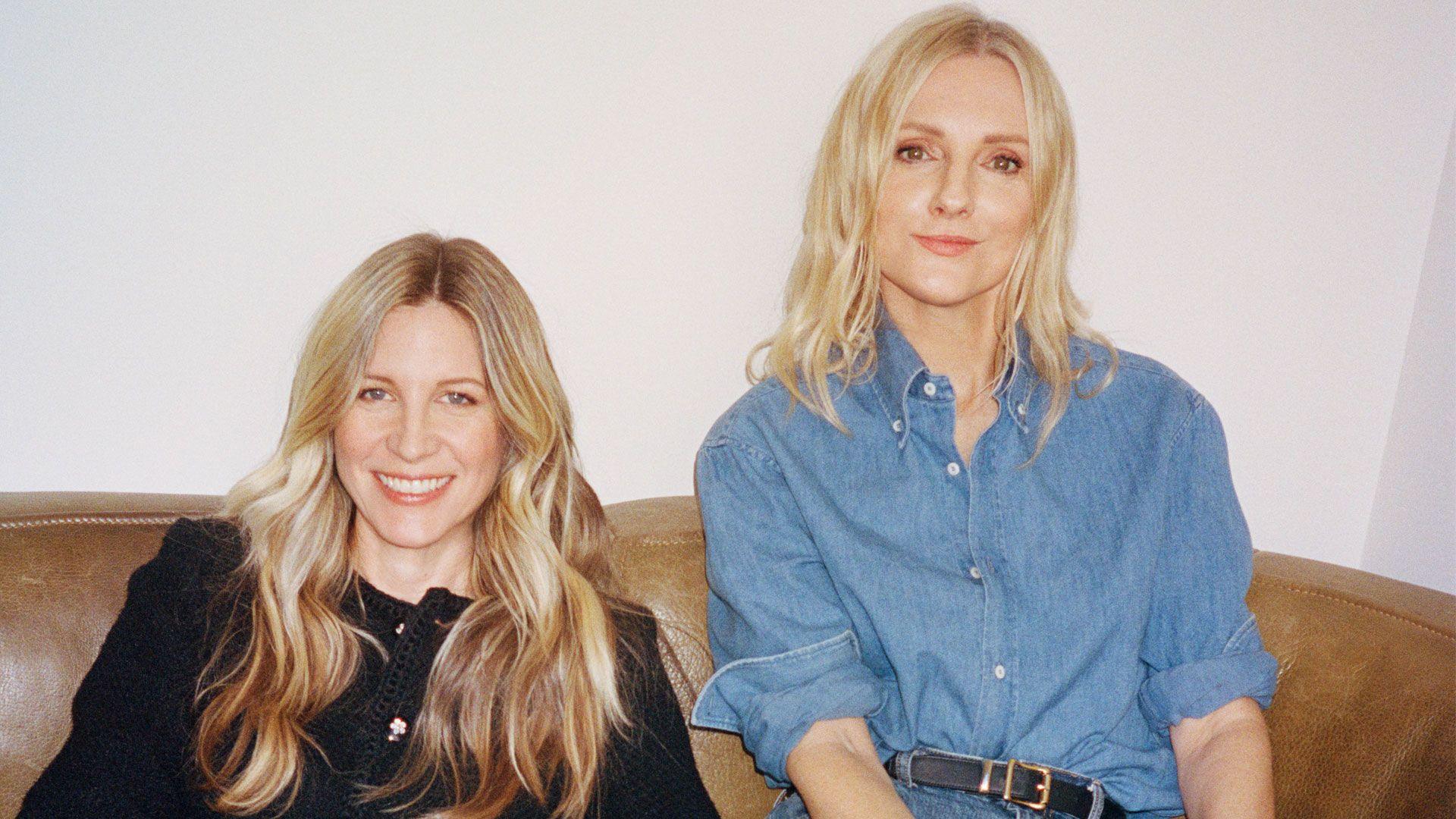 Two women with blonde hair smiling, one wearing a black jacket and the other a blue button up shirt.
