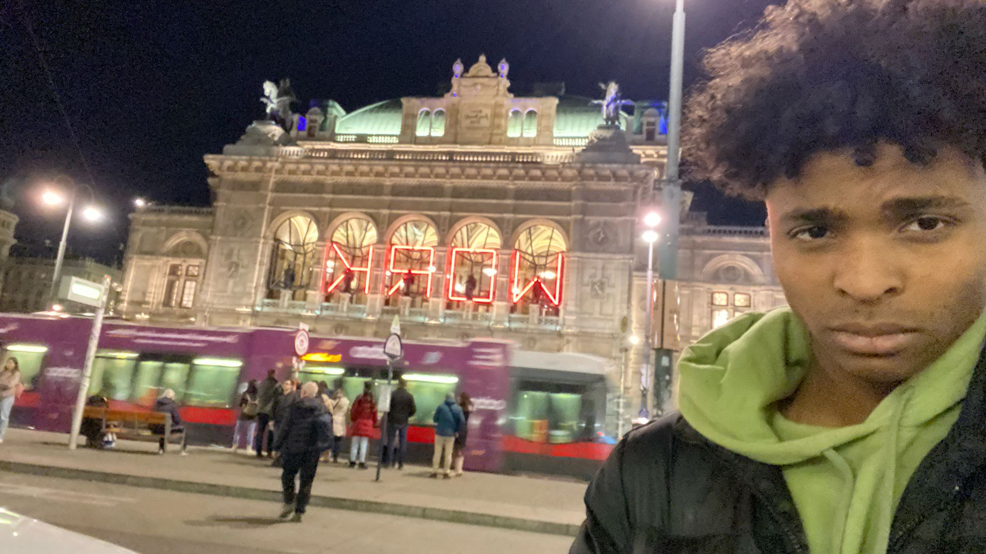 Luka stands next to a building in Vienna, with a tram going by behind him. He is looking at the camera. 