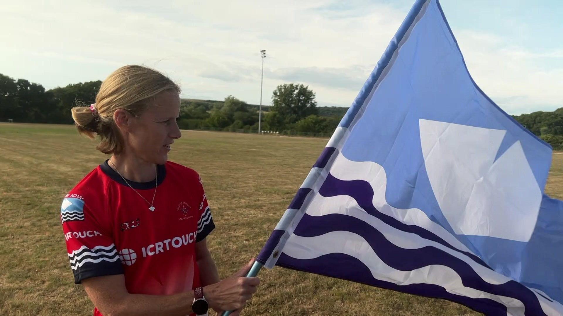 Sammie Mumford standing on a playing field, wearing a red Team Isle of Wight top and holding a blue Isle of Wight flag