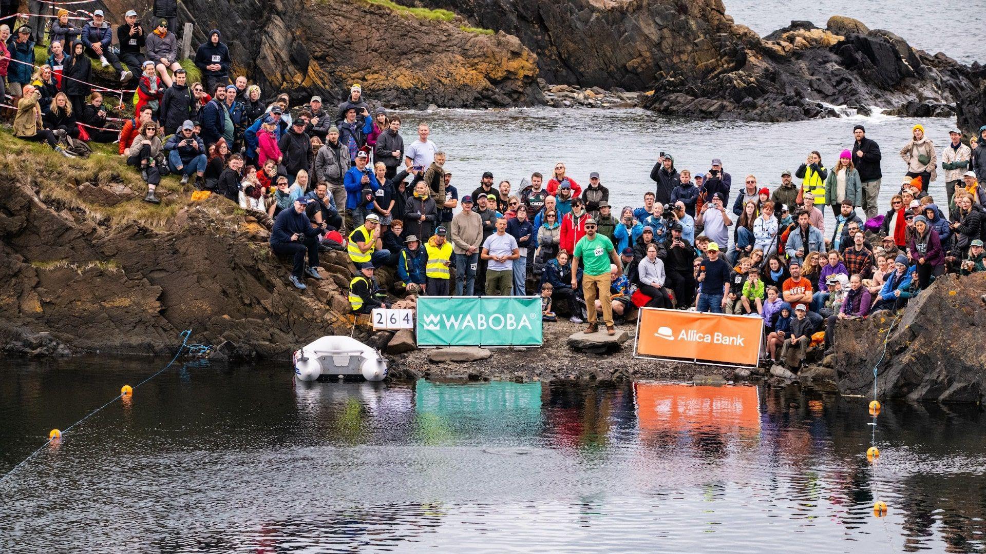 Jon Jennings wears a green top, beige trousers, a cap and sun glasses as he stands on the edge of a flooded quarry to skim a stone. Around him is a crowd of people. There are sponsor banners perched on the edge of the small rocky strip of land he is standing on. One says WABOBA, the other says Allica Bank. A small inflatable boat is moored next to the rocks. Water stretches out being the group.