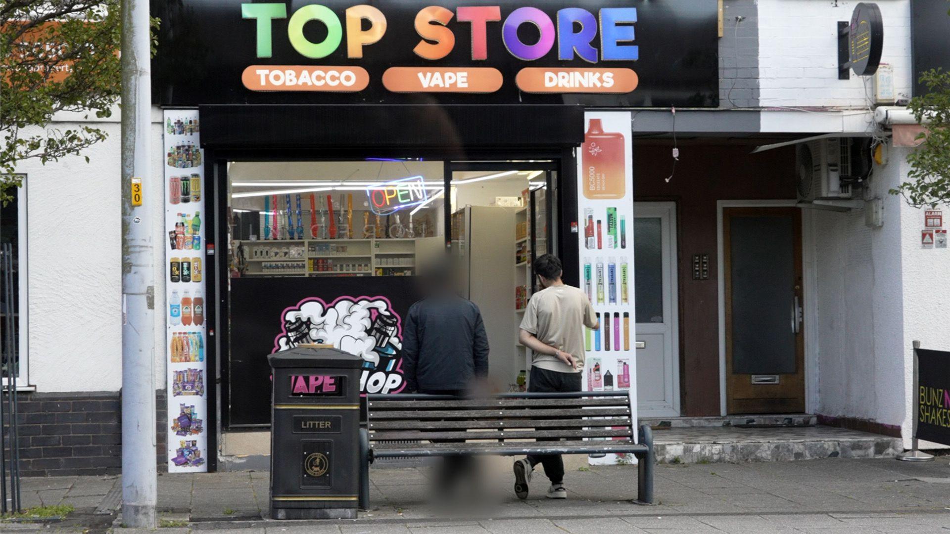 A photo of a mini-mart with a brightly coloured sign which reads Top Store. Outside are ten men with their backs to the camera, leaning against a bench. One is our undercover journalist, which is blurred to protect his identity. The other is the shopkeeper, Surchi.  