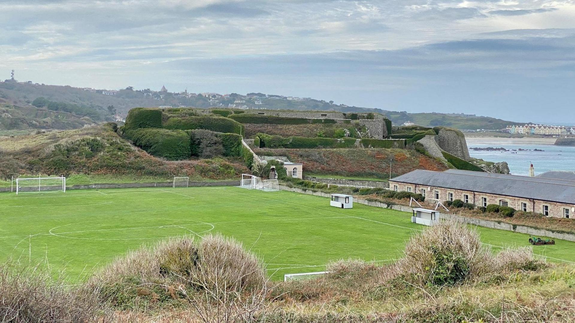 The Arsenal ground in Alderney. The football pitch is marked out with white lines. A long single storey building is to the right of the pitch. The sea is in the background.