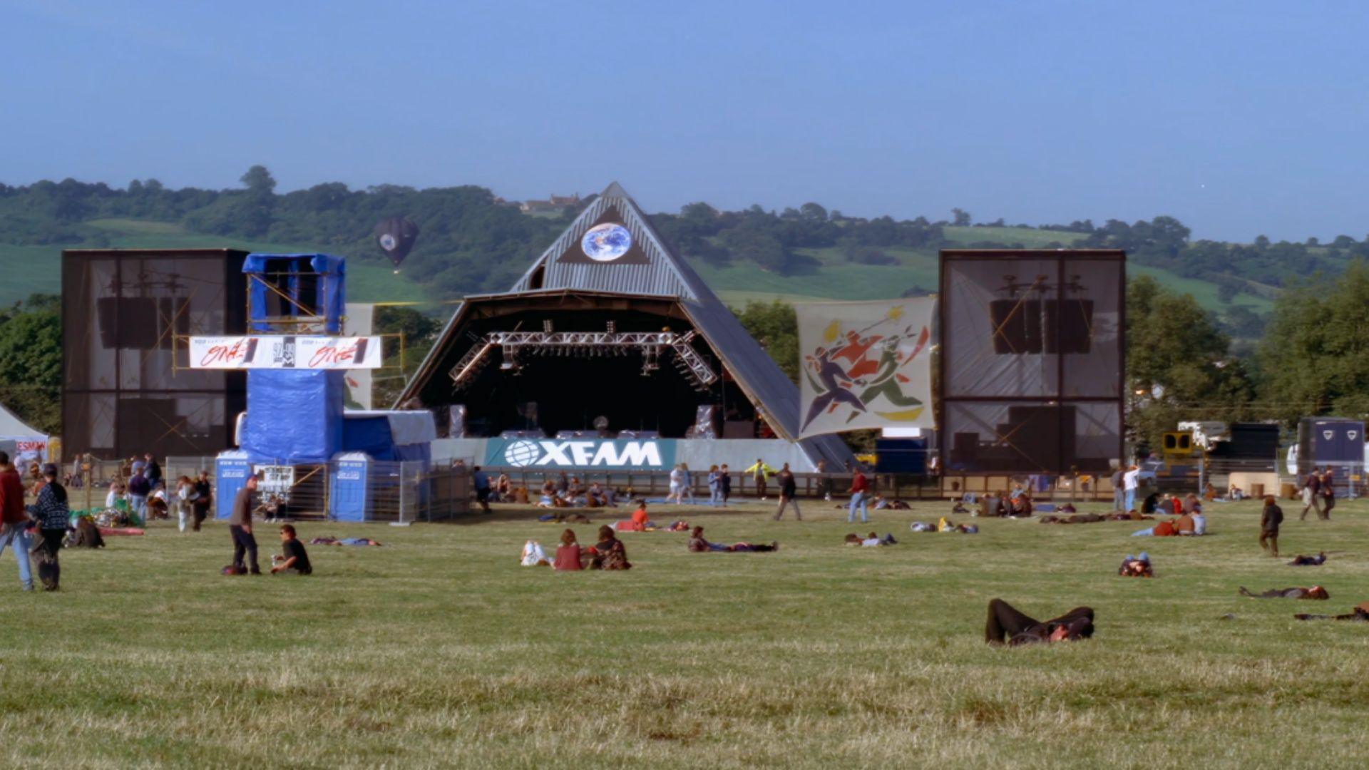 A vintage image of a field with the early Pyramid Stage in the background and a few scattered festivalgoers sitting on the grass. There is a blue sky and hills behind