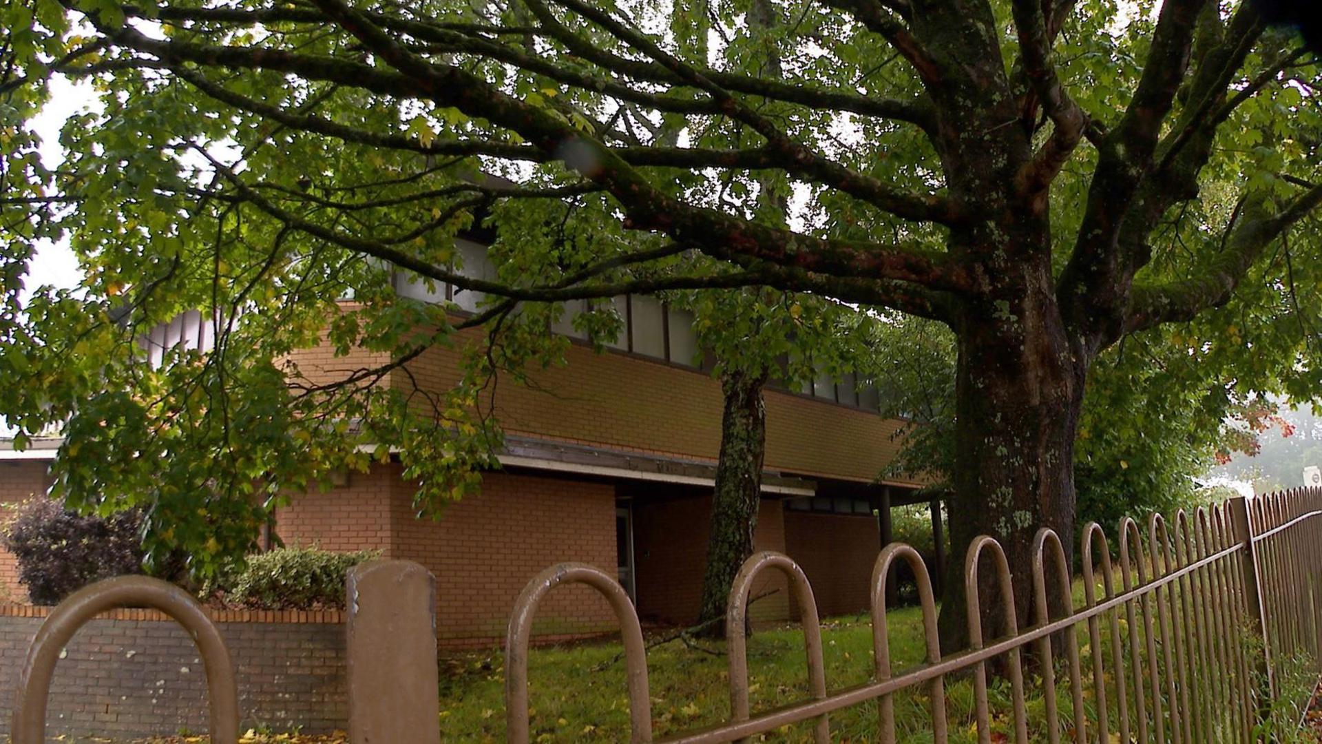 The old register office. The brick, two story office building. The view is over a brown metal fence  showing some grass and a tall tree in the grounds. 