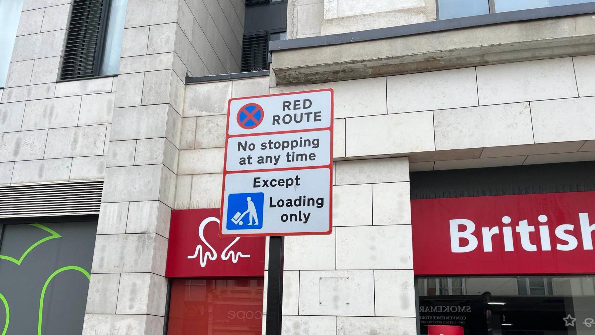 A white road sign with red borders on a street in Brighton, it reads "red route, no stopping at any time, except loading only"