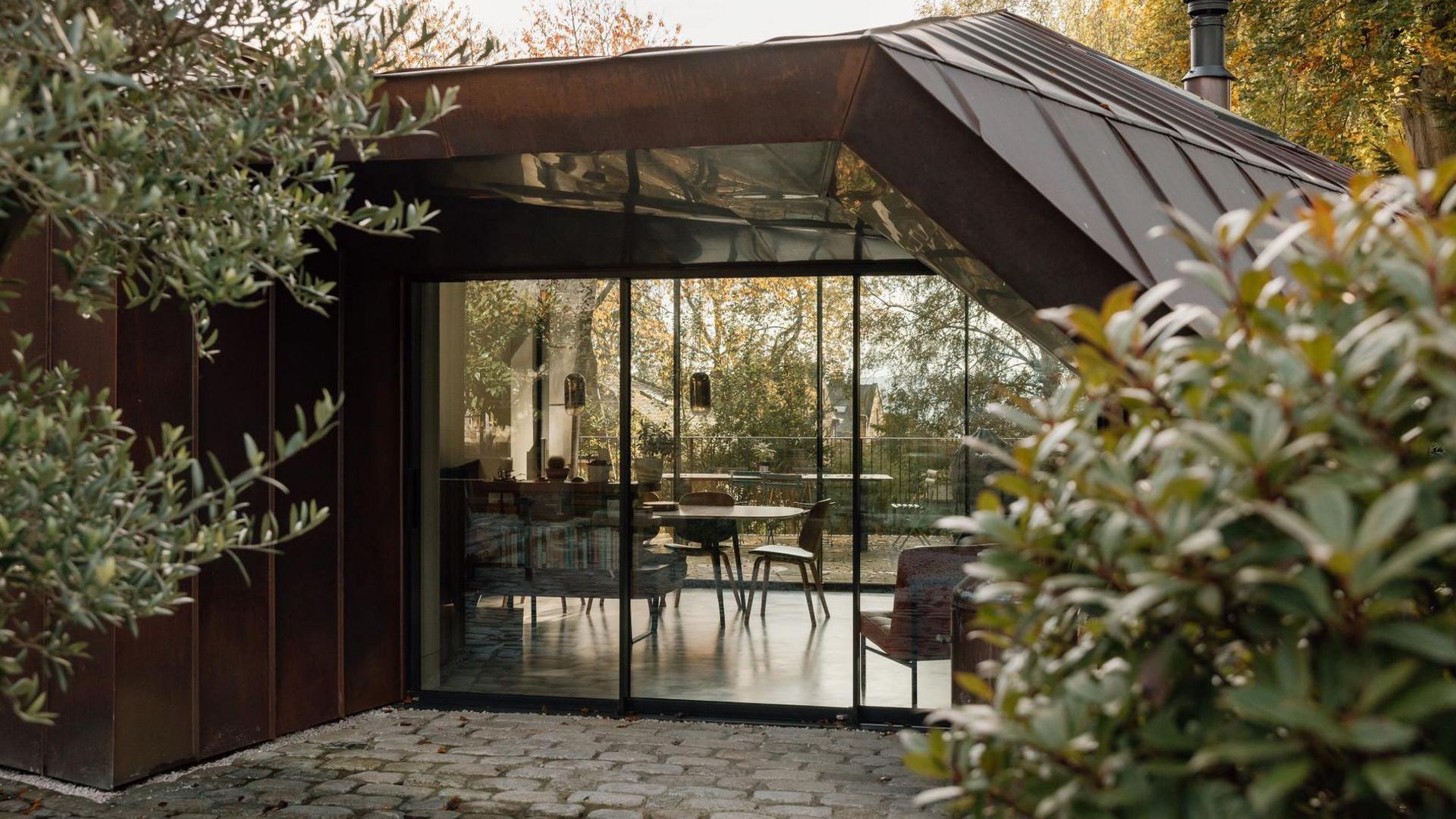 The image shows one of the rooms behind a glass front. A dining table, chairs and lamps can be seen through the floor-length windows.