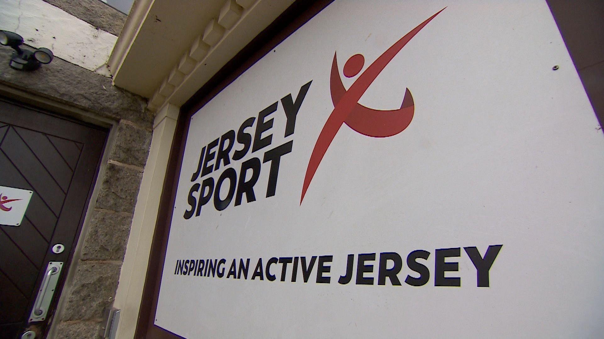 A large white sign, with black writing, which says: "Jersey Sport, inspiring an active Jersey." It has a red logo in the shape of an 'x' with a circle at the top. The sign is on a wall, on the outside of a building, next to a dark brown, wooden door.