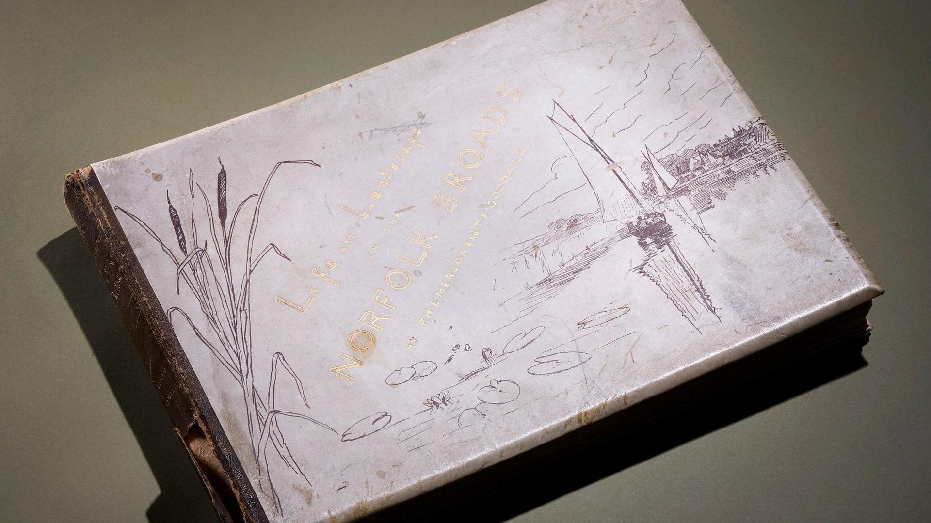 View of the cover of 'Life and Landscape on the Norfolk Broads' by Peter Henry Emerson. The title is written in gold ink and the white paper has drawings of reeds and water symbolising the Norfolk Broads.