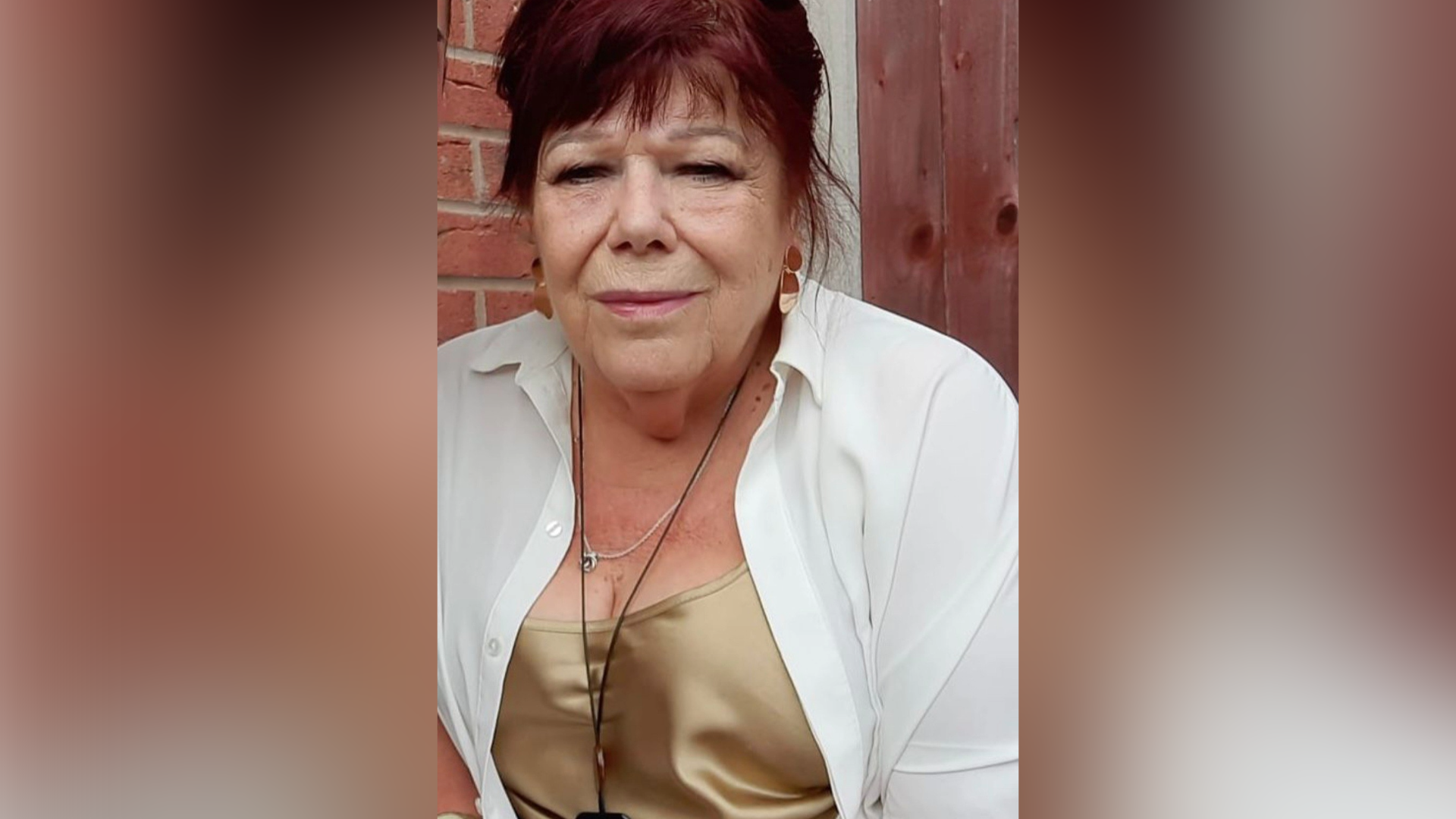 A woman with red hair wearing a white buttoned shirt and shiny gold top is pictured with a brick wall behind her and a wooden fence over her left shoulder.