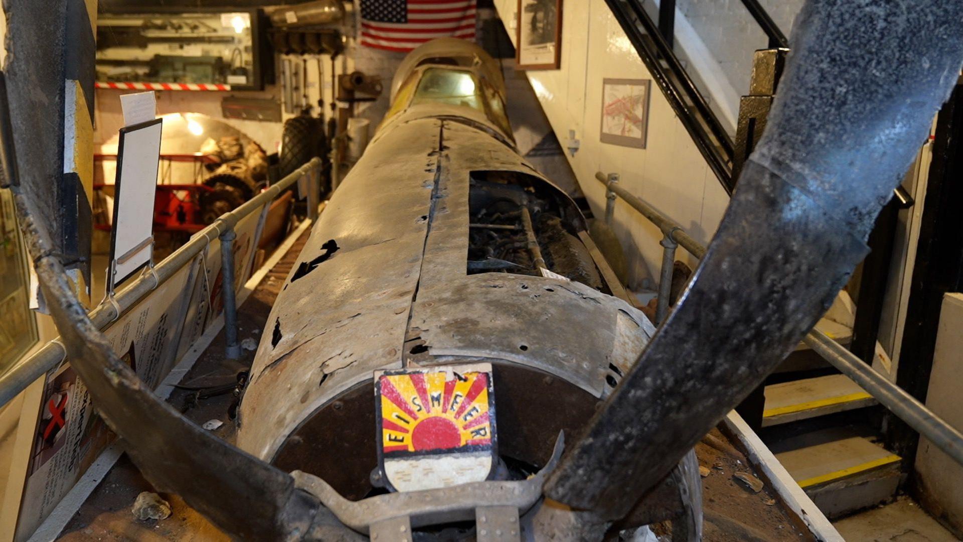 An aircraft wreck with twisted faded metal is on display in a museum. Behind Little Zippie, the American flag is flying and there are railings preventing people from touching the exhibit.