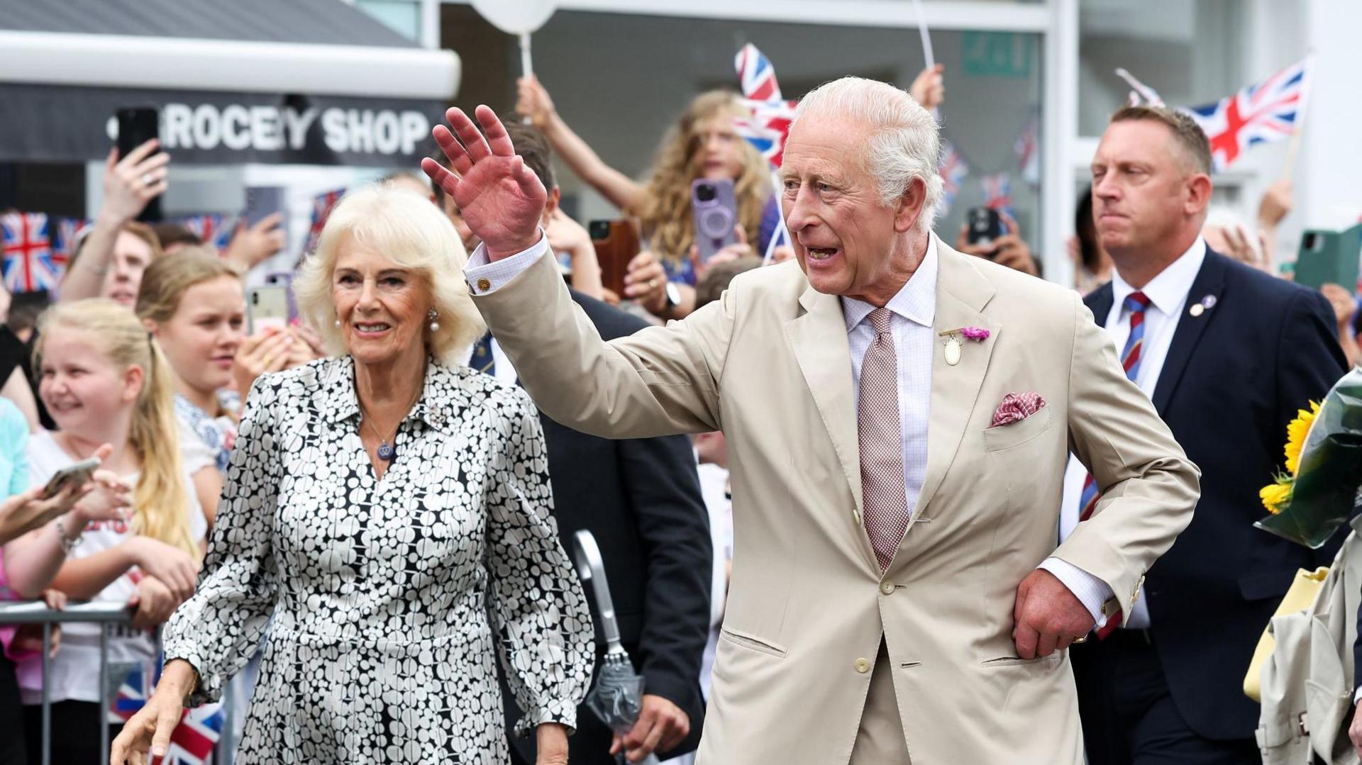 The King waves as he walks along Newmarket High Street. The Queen walks beside him. They are both smiling.
