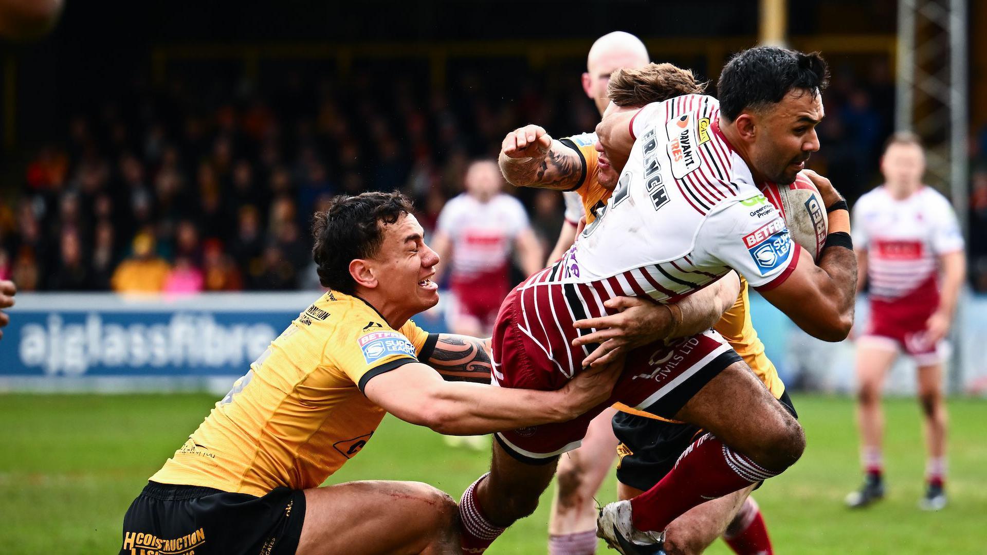 Wigan's Bevan French (right) has the ball in his right hand as two Castleford players including Daejarn Asi (left) try to tackle him