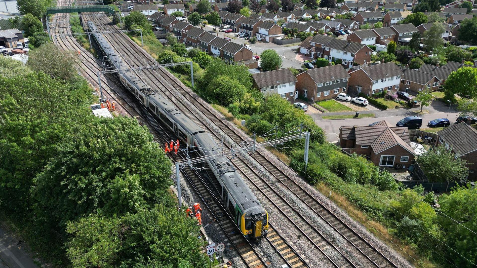 An aerial view of the whole train sitting slightly skewed on the tracks, with silver gantries overhead. There is a housing estate to the right with many semis and terraces, and bushes to the left. Several workers in orange high vis clothes mill around.