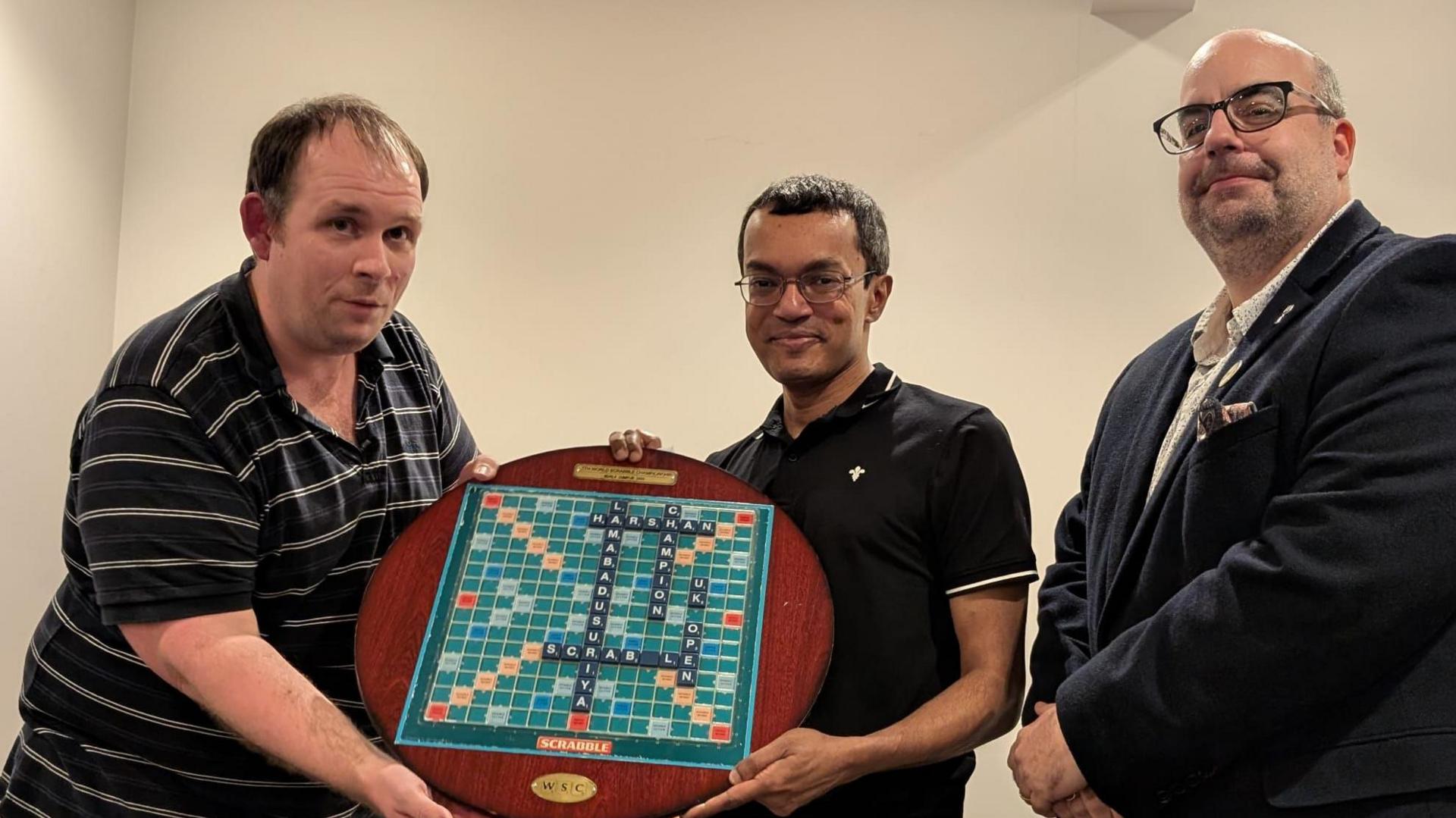 James Burley hands Harshan Lamabadusuriya his award, which is a large wooden circular plinth with a scrabble board on it. On the board, spelt and interlocked in black tiles, is 'Harshan', 'Lamabadusuriya', 'UK', 'Open', 'Scrabble', and 'Championships'. Holiday Inn sales and marketing manager David Berry stands next to them, his arms crossed in front of him. James has short brown hair and wears a black sports shirt with white stripes. Harshan has short black hair, glasses, and a black sports shirt. David is bald on top with short grey hair on the sides, and wears a suit with a collared shirt and no tie.