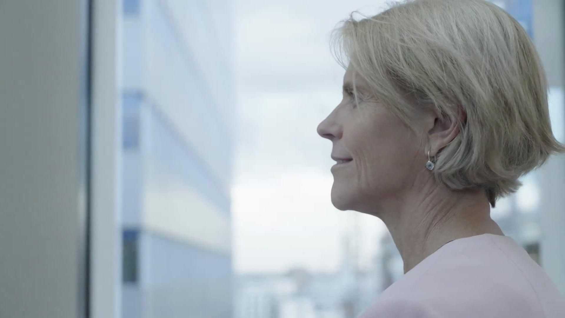 Meagen Burnett, a woman with short blonde hair wearing a light pink top, looking out of a window in a high rise office building