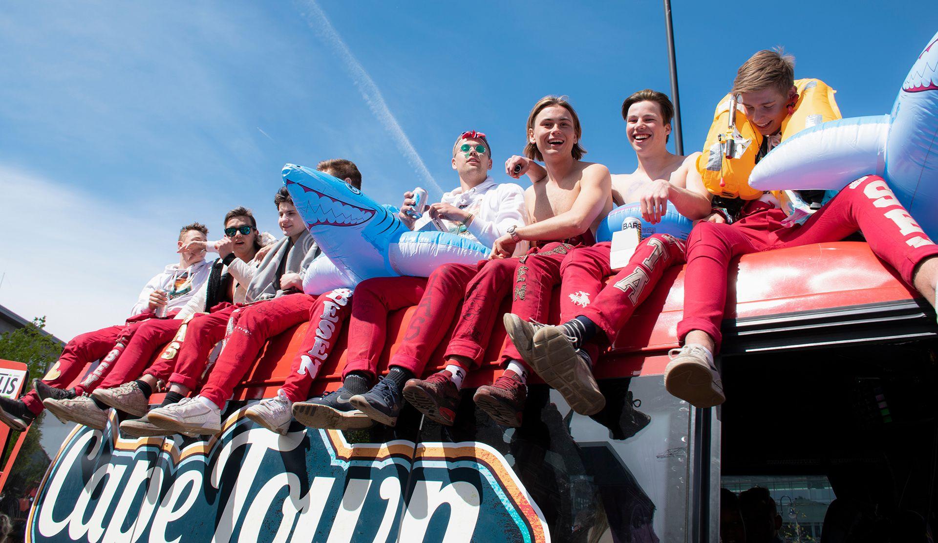 A group of school-leavers in red overalls sit on a red russebuss smiling and drinking and with inflatable pool toys against a clear blue sky, with the words 'Cape Town' visible on the side of the bus, in Arendal on 17 May 2019.