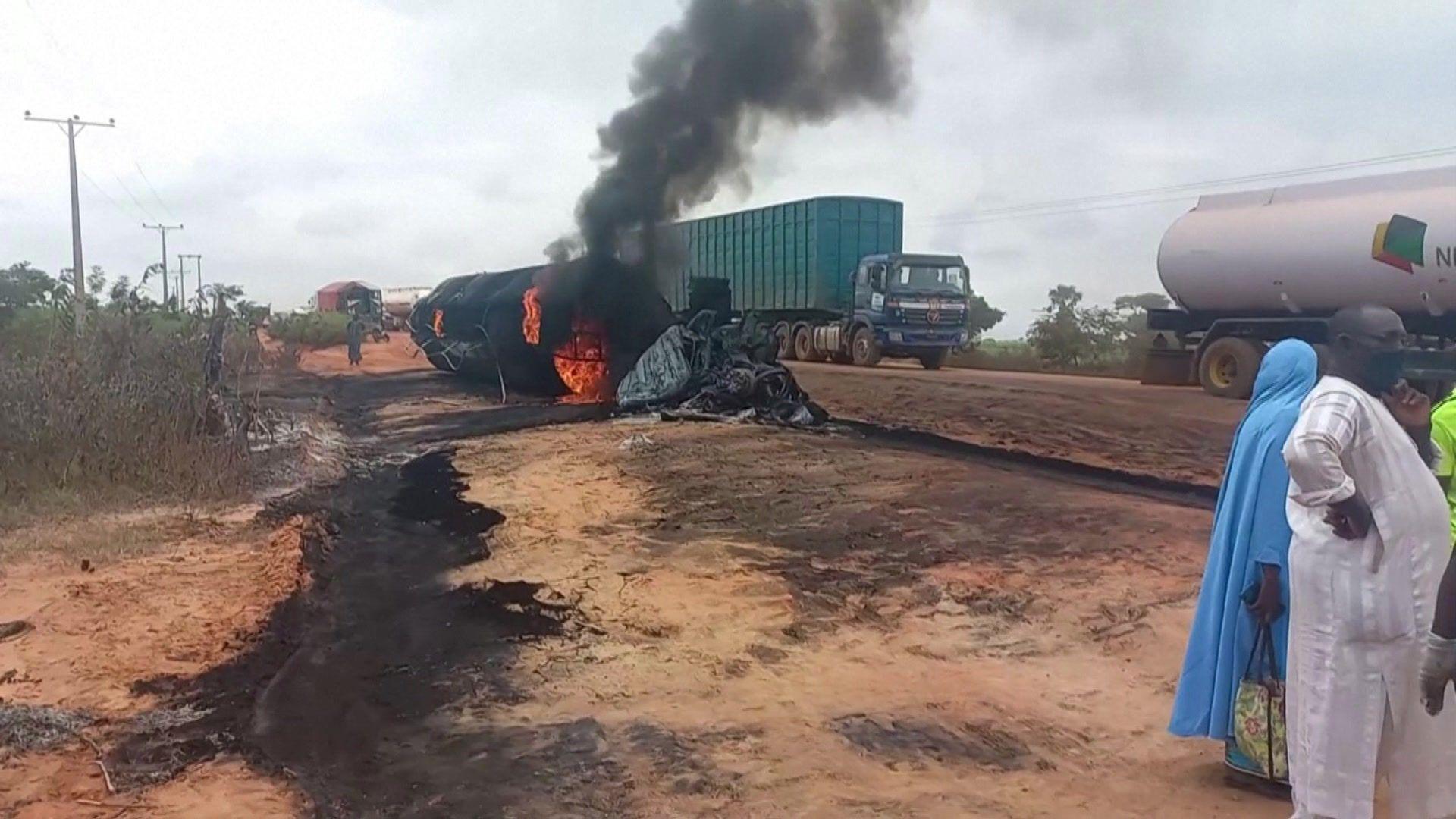 A flaming burnt-out oil tanker lies at the side of the road in the state of Niger. Its shell has been completely blackened. On the nearby road, which is not paved, a lorry and tanker can still be seen. There are burn marks on the ground and some people can be seen in the near ground. 