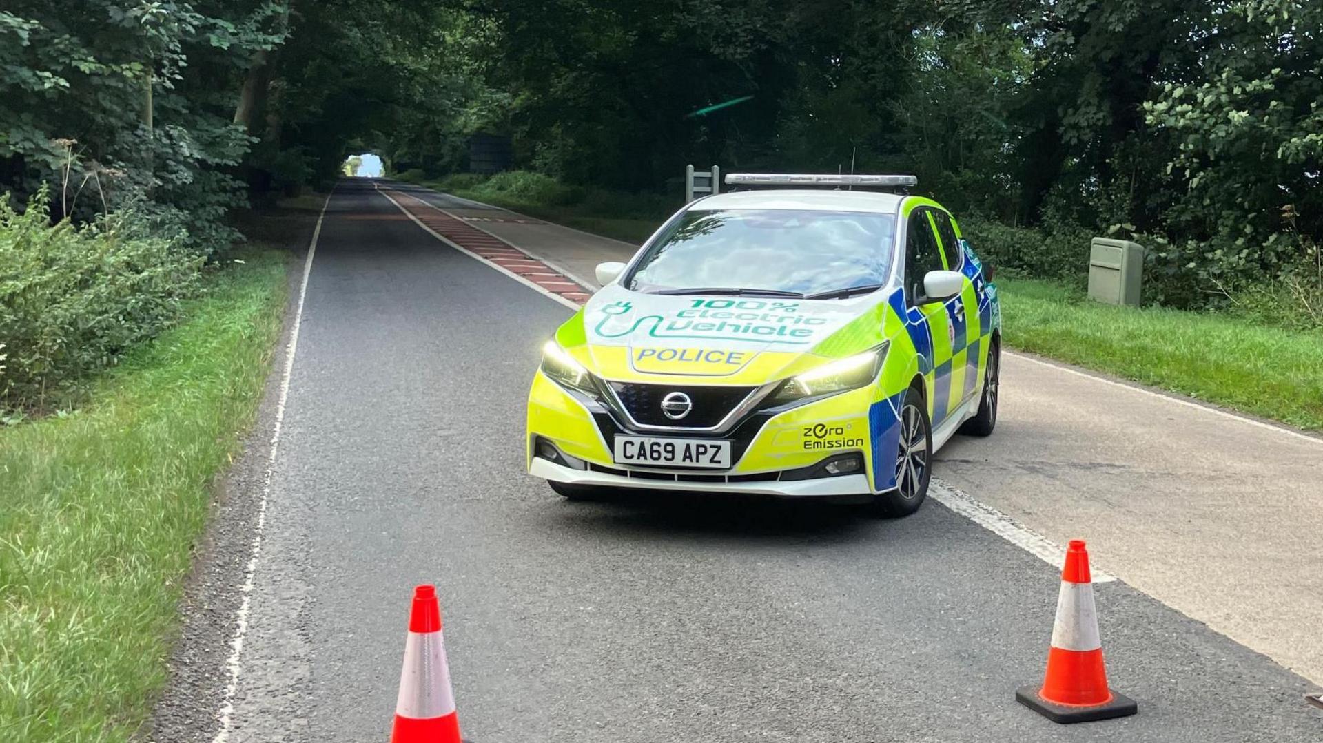 A police car parked in the middle of the road with two orange cones placed across the road to block oncoming traffic.