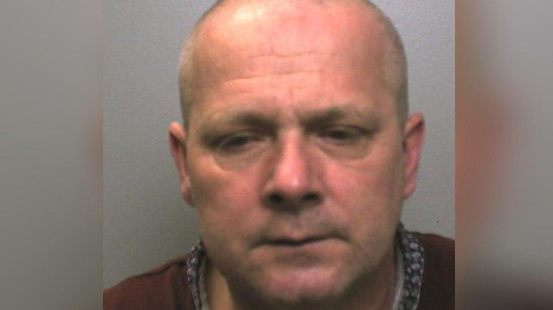 A mugshot of a bald man wearing a red jumper. He is looking down in a crestfallen manner.