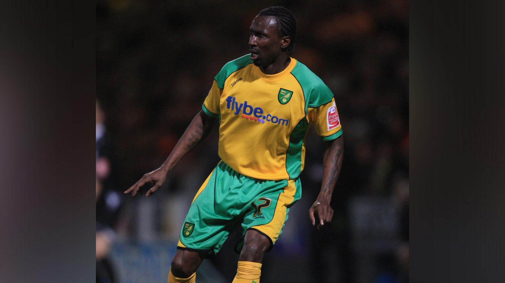 A man in a yellow and green football strip with "Flybe.com" on the front looks to the left. The crowd in the football stadium behind him is blurred.
