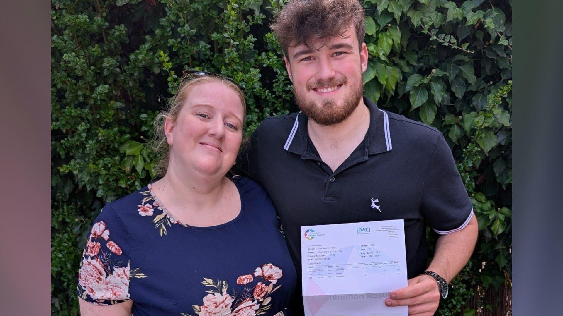 Amber wearing a navy blue dress with flowers standing next to Ollie wearing a blue T-shirt and holding his results in the hand