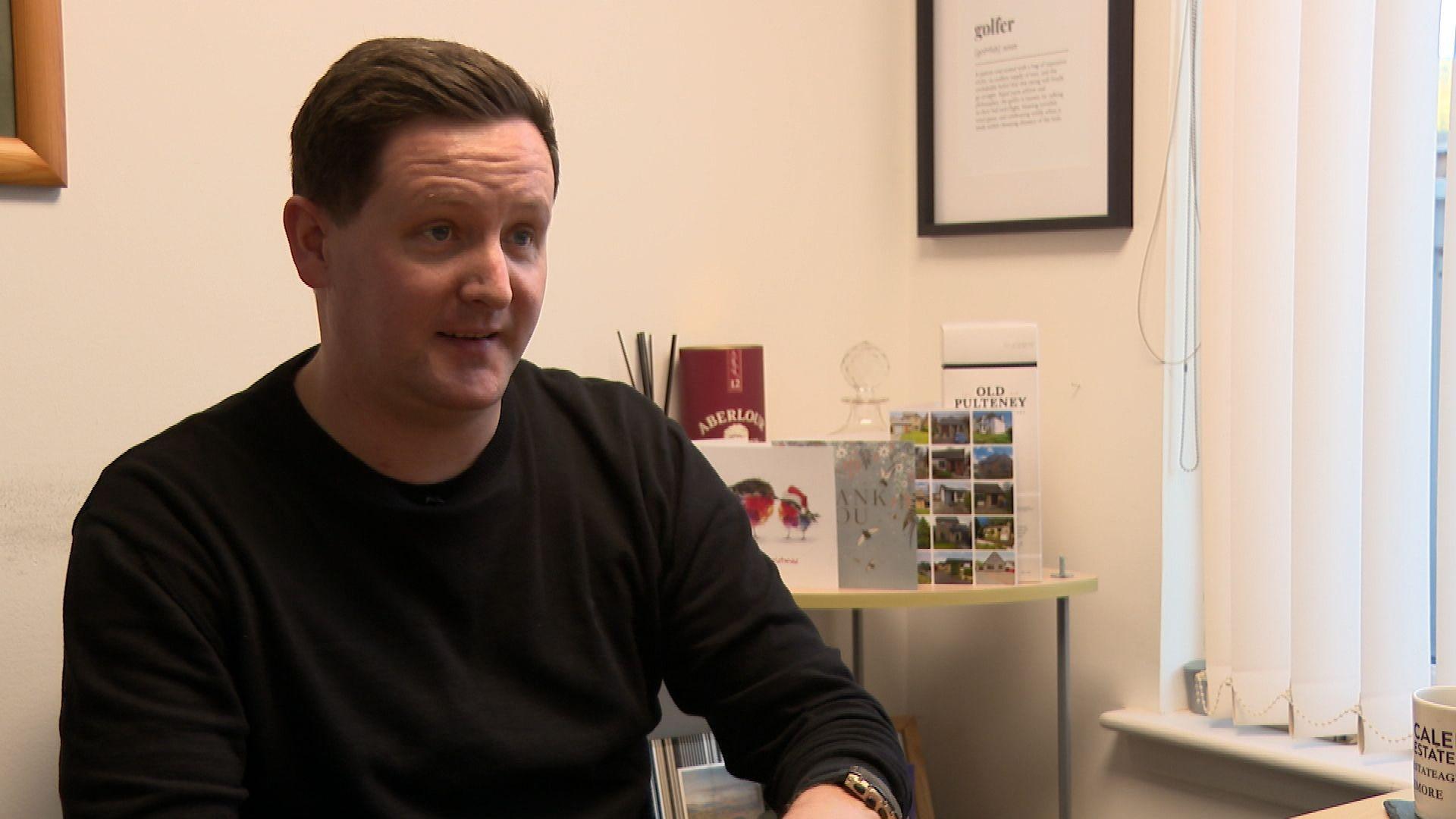 Darren McConnell bhon chompanaidh Caledonia Estate Agency ann an geansaidh dubh, na shuidhe faisg air uinneig