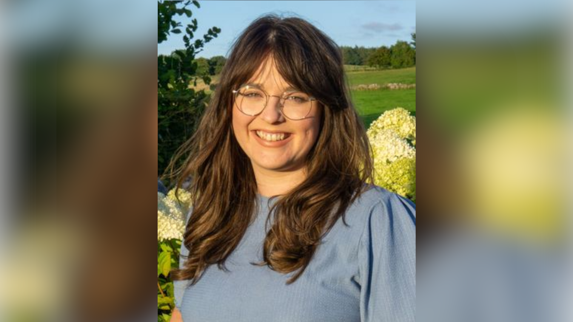 A picture of Dr Emily Haythornthwaite. She is smiling at the camera while standing outdoors with a green field and flowers pictured behind her. She has long brown hair with bangs. She is wearing a blue top and glasses.