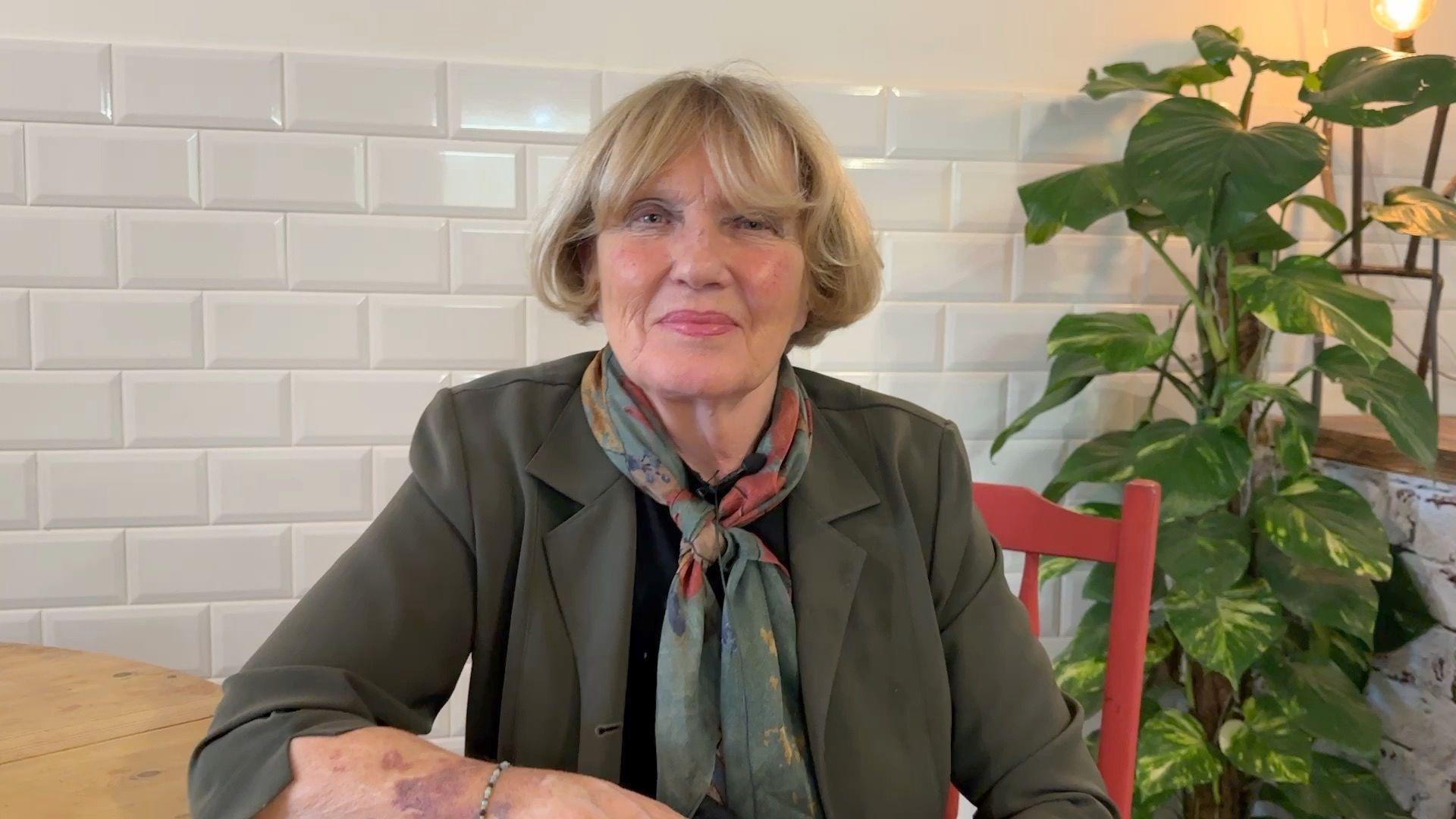 Denise McKay with blonde hair, wearing an olive blazer and a pashmina. She is sitting at a wooden table on a red chair looking at the camera.