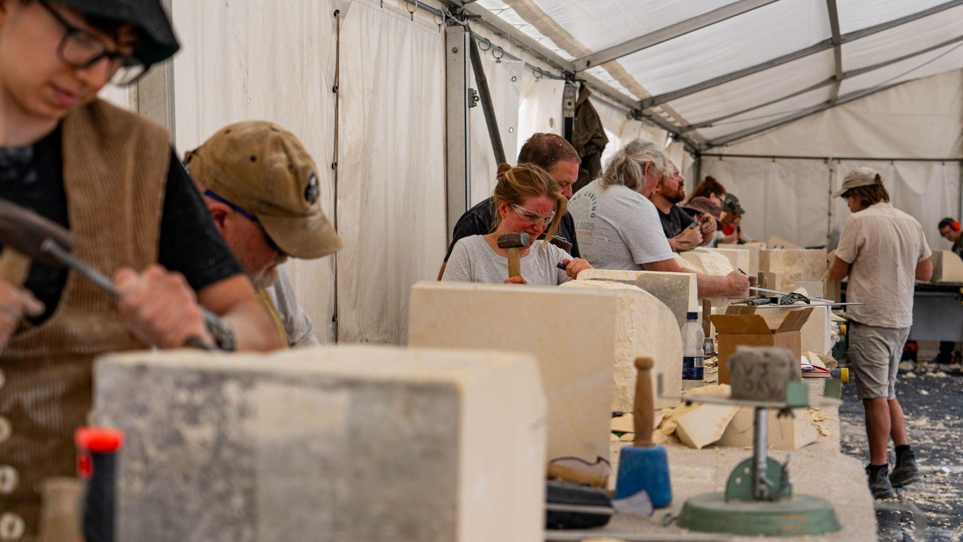 People in a line carving pieces of stone, some of them wearing protective masks and goggles