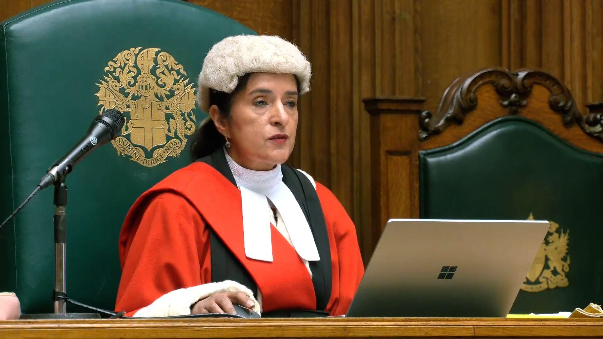 A female judge dressed in red robes and a wig. She has dark hair, tied back and is sat at a wooden desk with a Microsoft laptop in front of her.