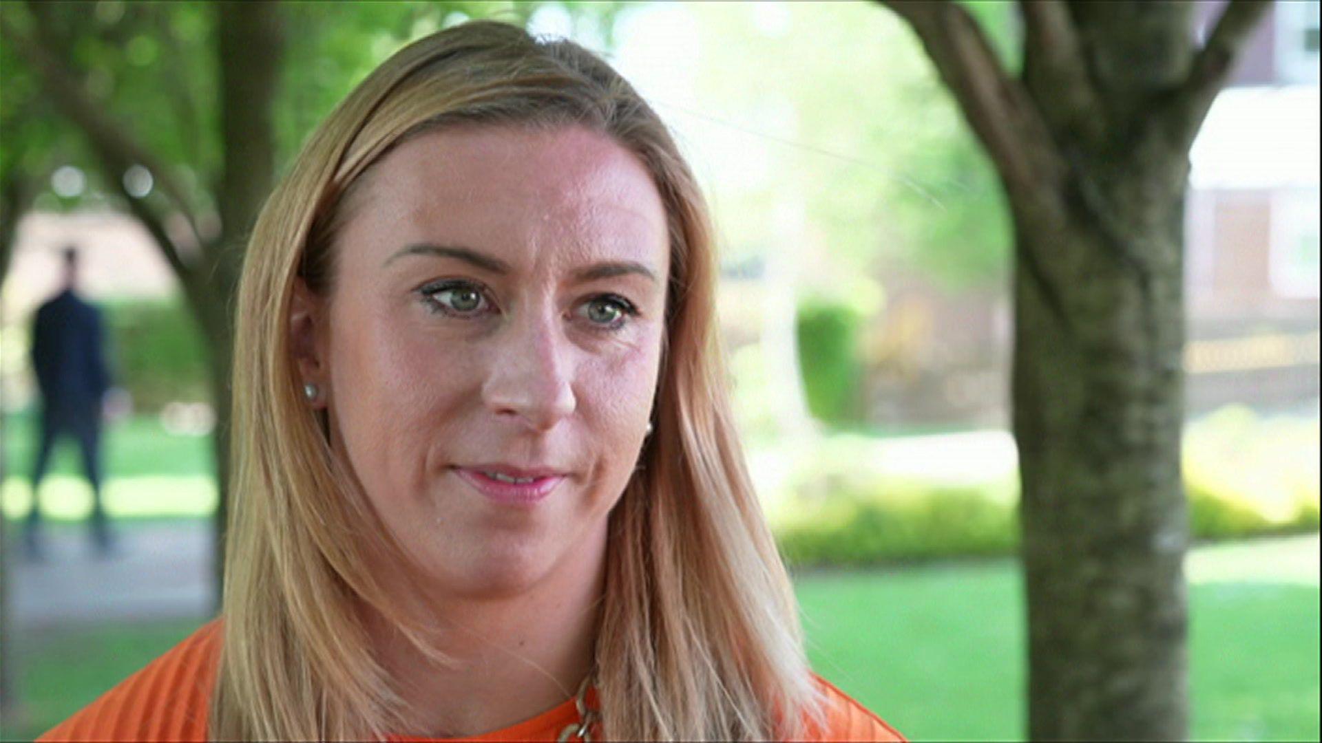 A woman wearing an orange top with blonde hair sitting infront of a blurry, glassy background