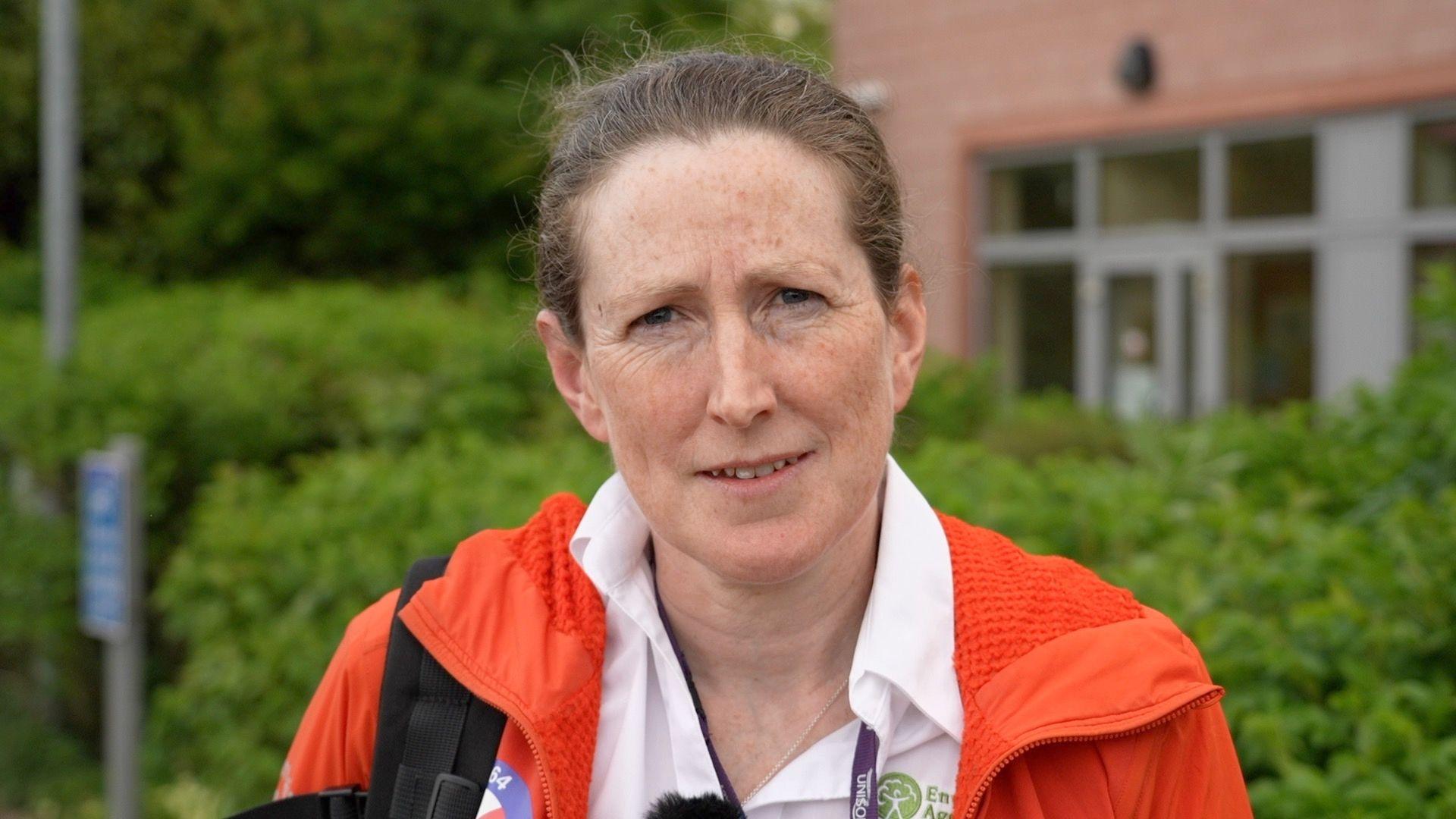 Amy Heys looks at the camera. She has brown hair pulled back in a pony tail, is wearing a white shirt with a green Environment Agency logo on the breast and an orange mountain rescue jacket. Behind her is a green hedge